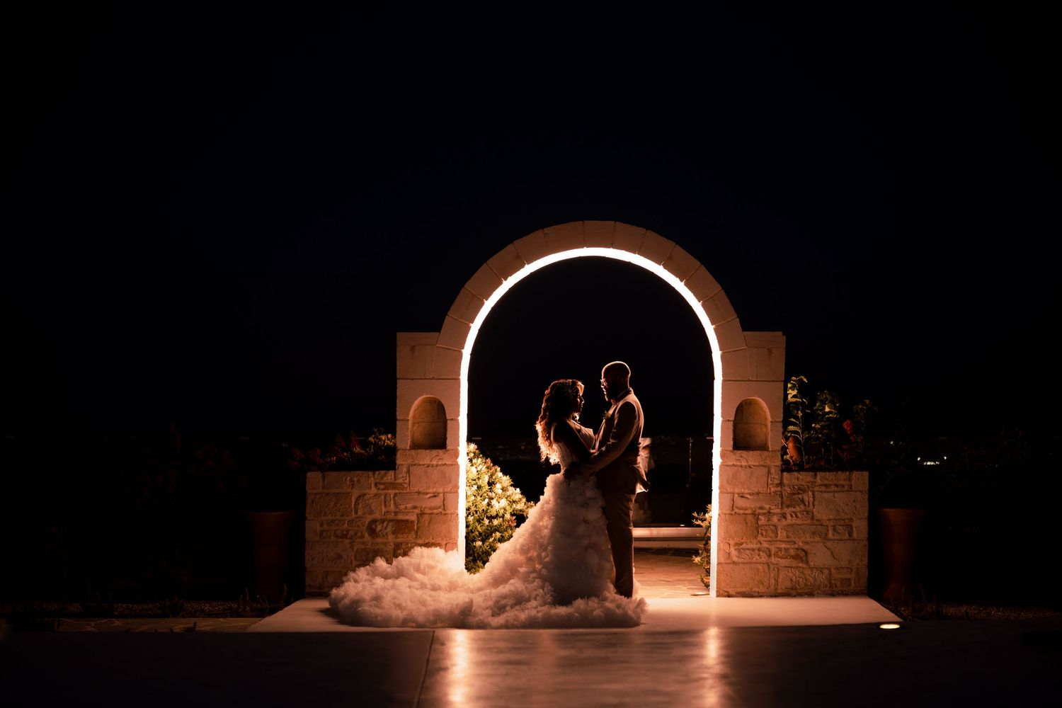 A silhouetted wedding couple stands in an illuminated stone archway at night.