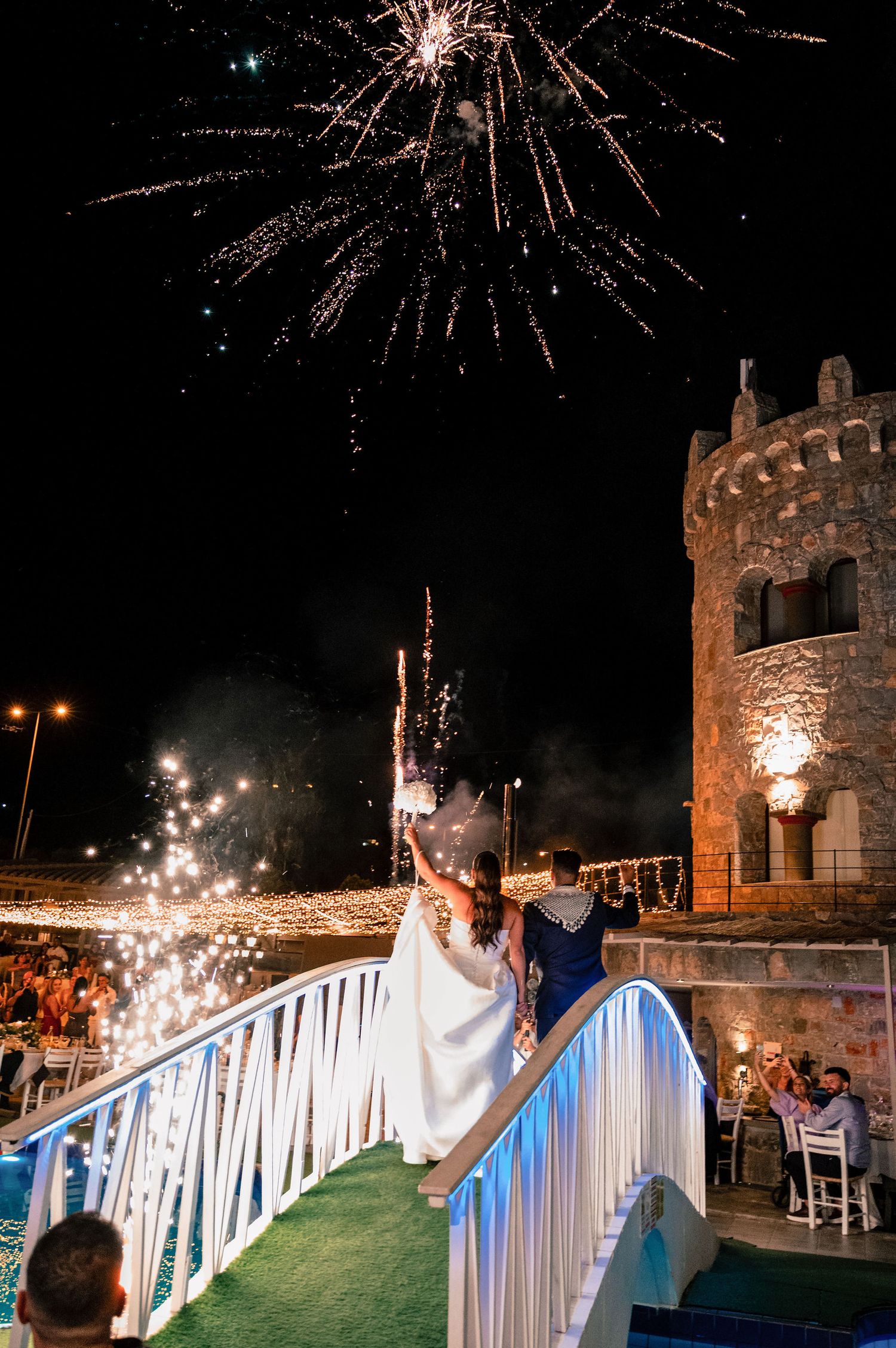 Fireworks light up the night sky over a stone castle structure with white and blue decorative railings.