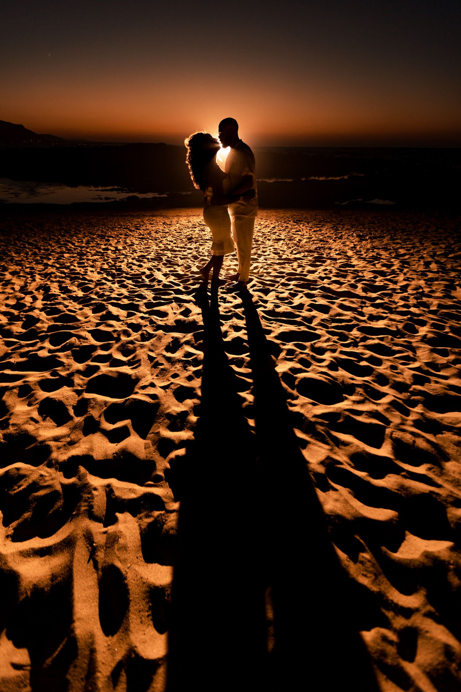 Silhouetted couple standing on textured beach sand during dramatic sunset.