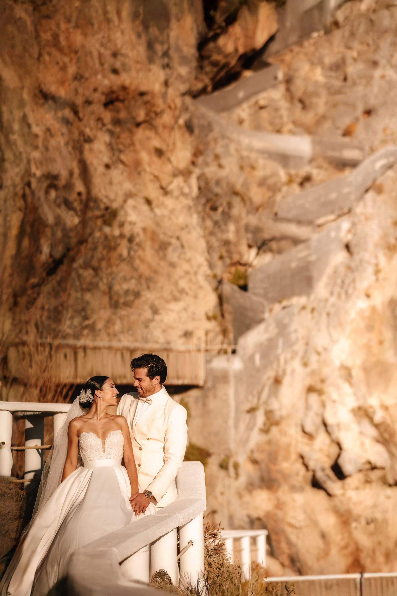A romantic wedding couple poses against a rustic stone wall backdrop in warm sepia tones.