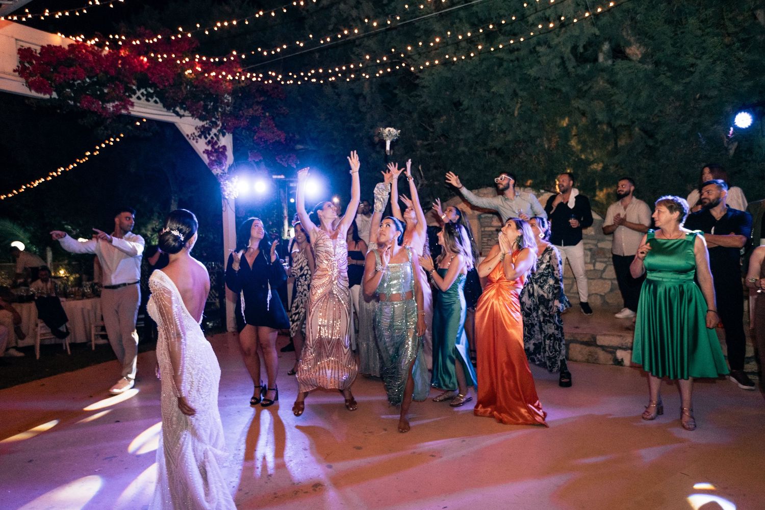 Colorfully dressed wedding guests dancing on outdoor reception dance floor under string lights.