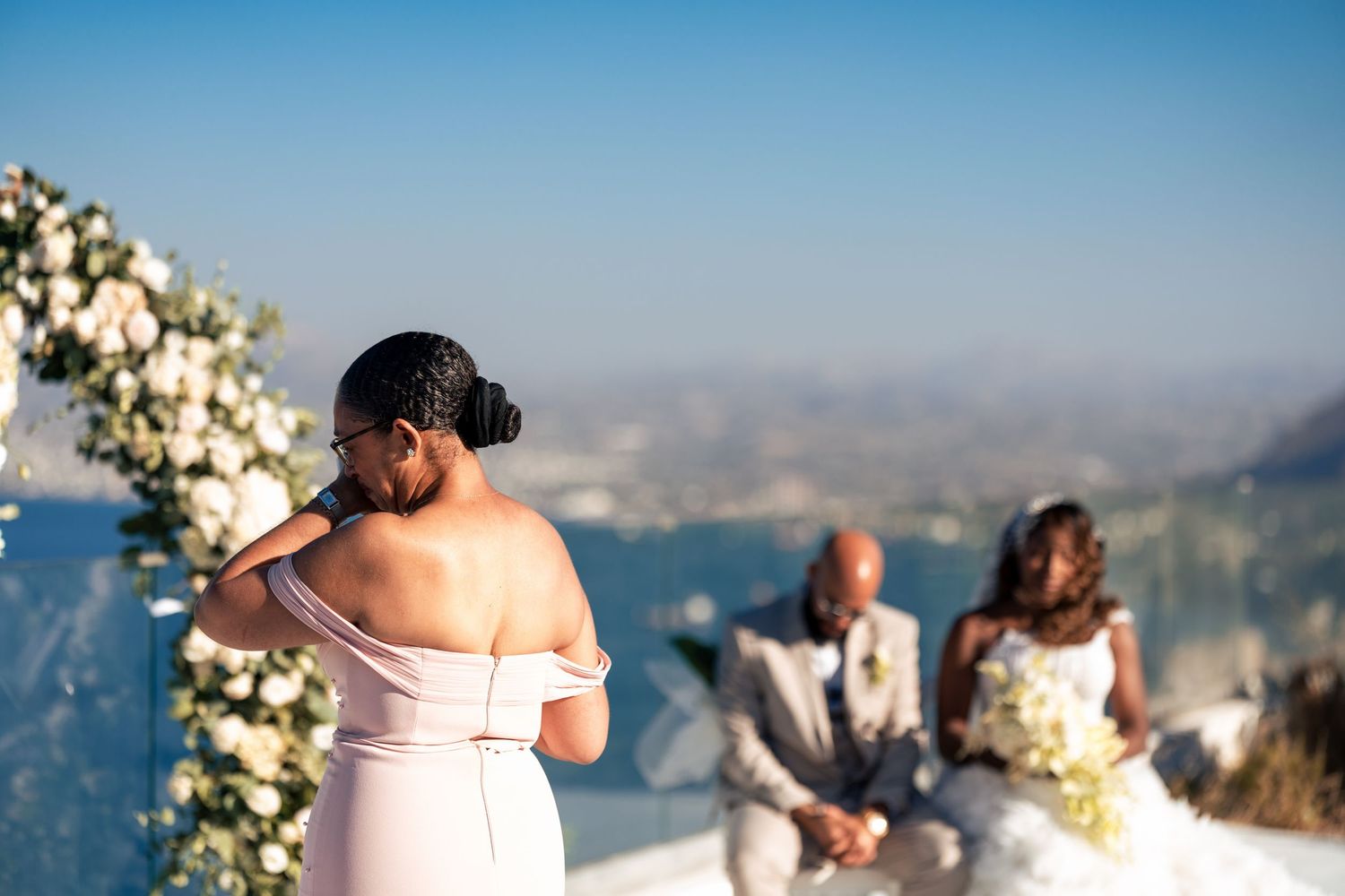 Seaside wedding ceremony with white floral decorations against blue sky.
