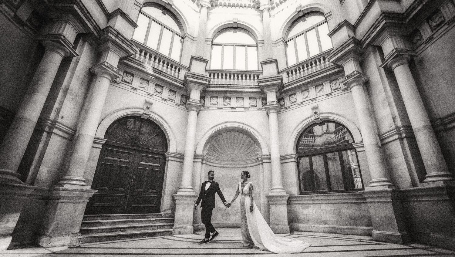 Black and white photo of couple holding hands in grand domed classical building.