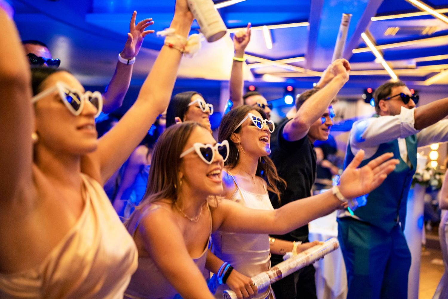 Wedding guests dance with arms raised under blue lighting at reception party.