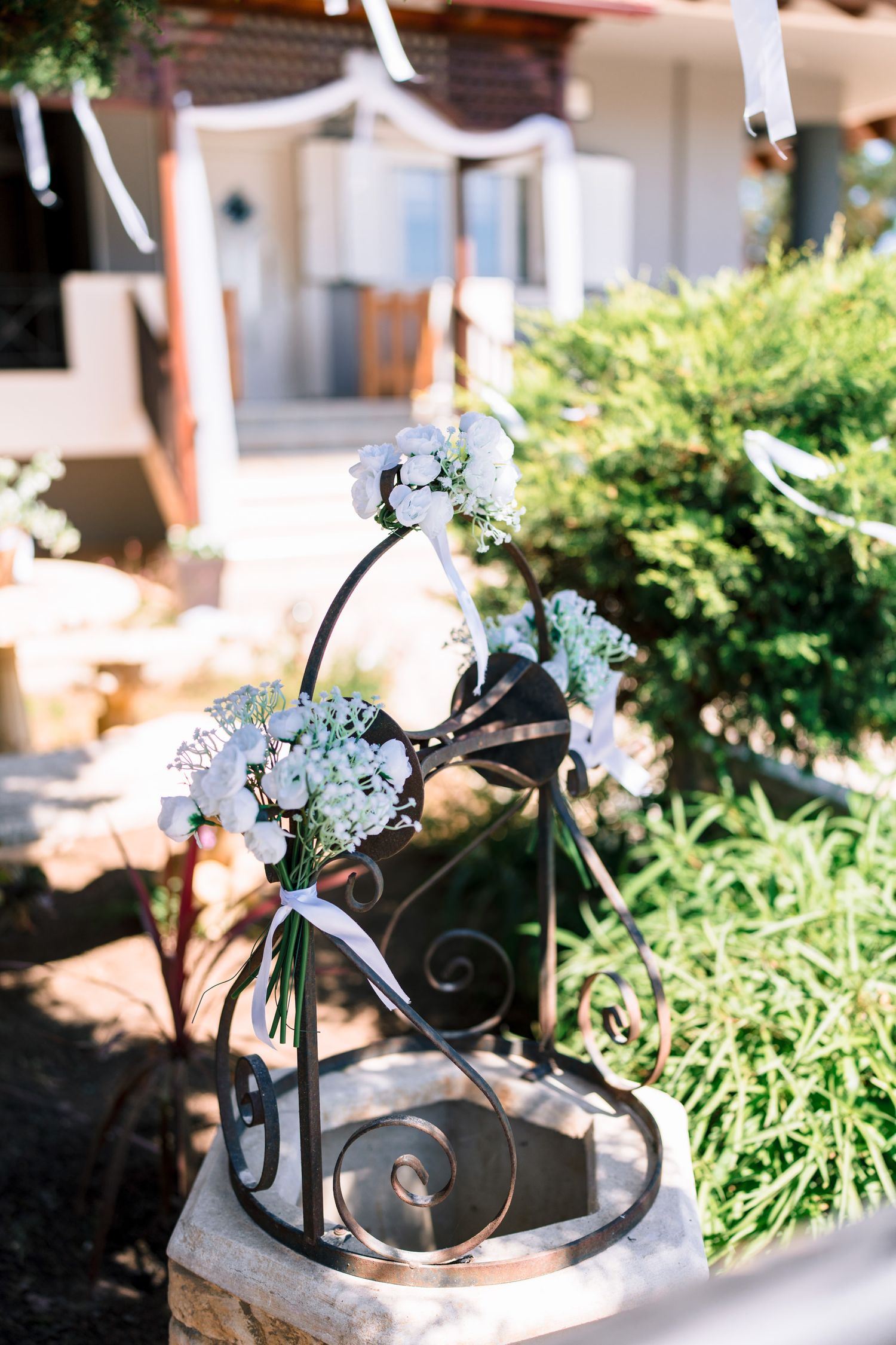 Decorative black metal wedding ring holder with small white flowers and greenery against a garden background.