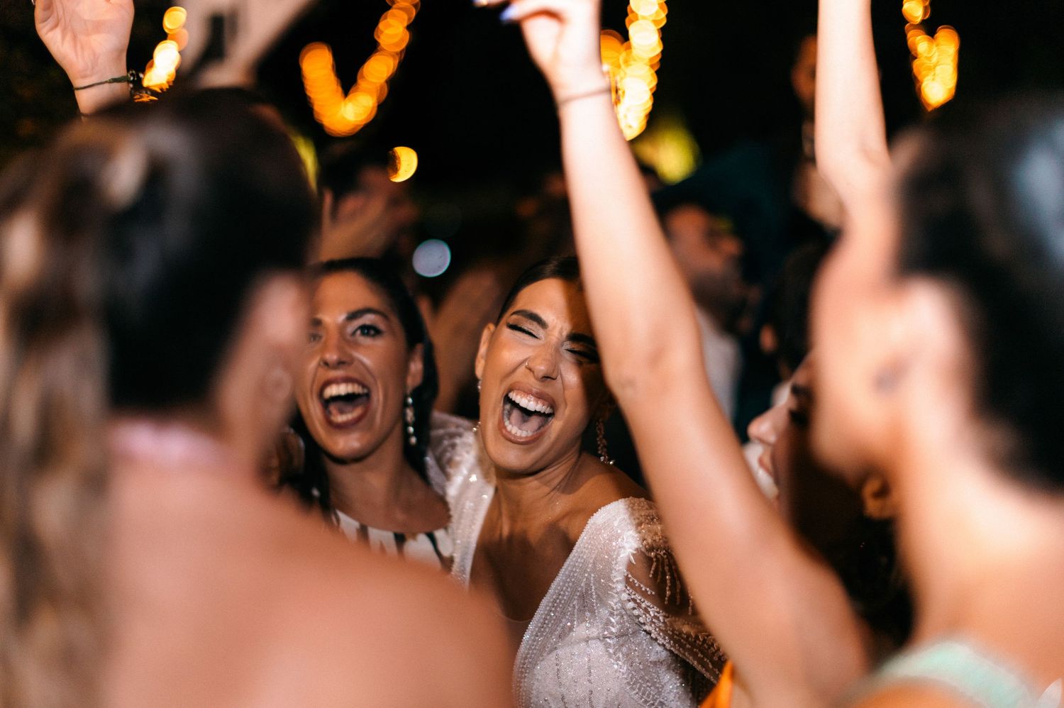 Wedding reception guests dancing and celebrating with arms raised under evening lights.