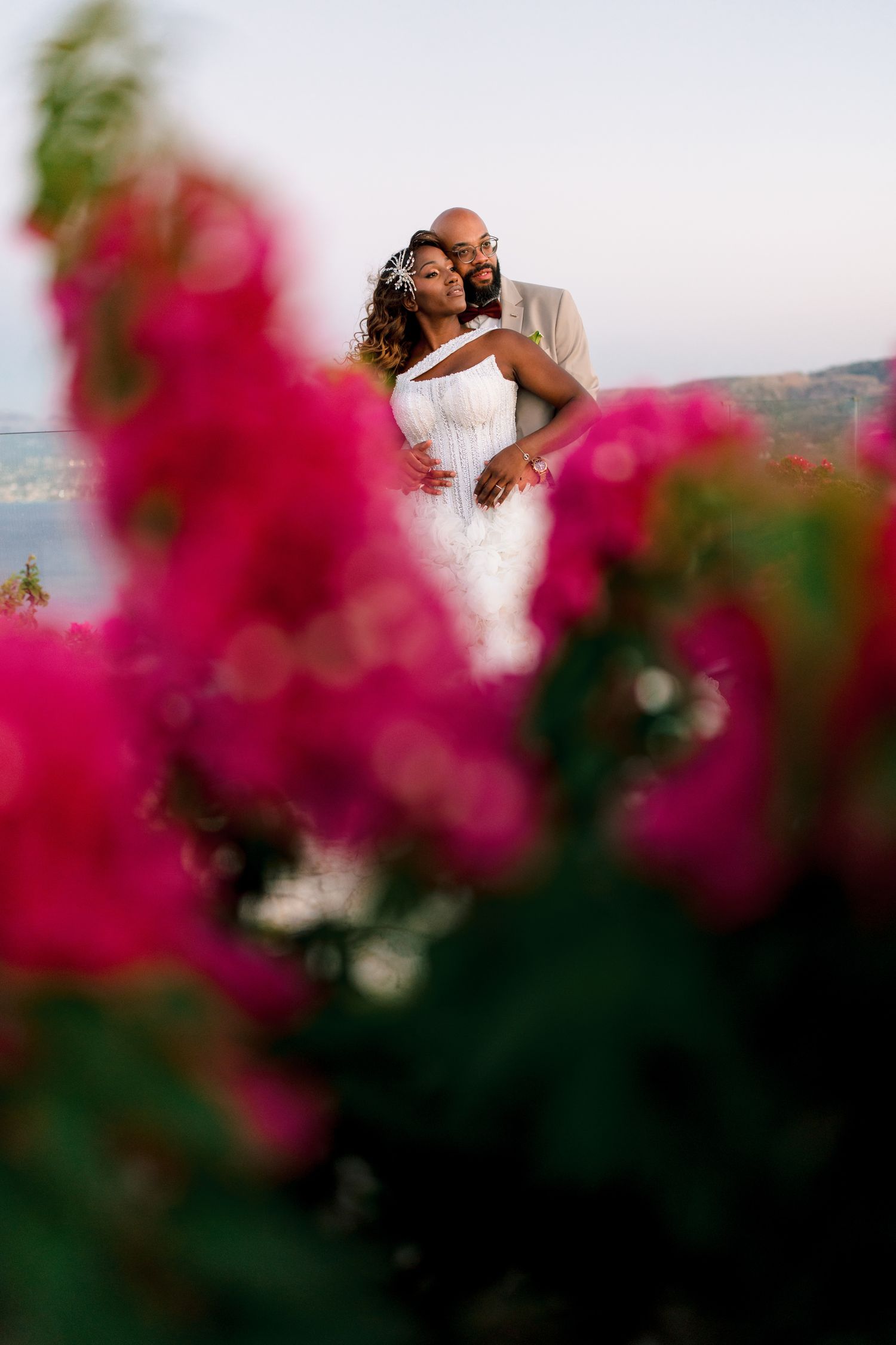 A romantic wedding photo framed by vibrant pink bougainvillea flowers with a coastal backdrop.