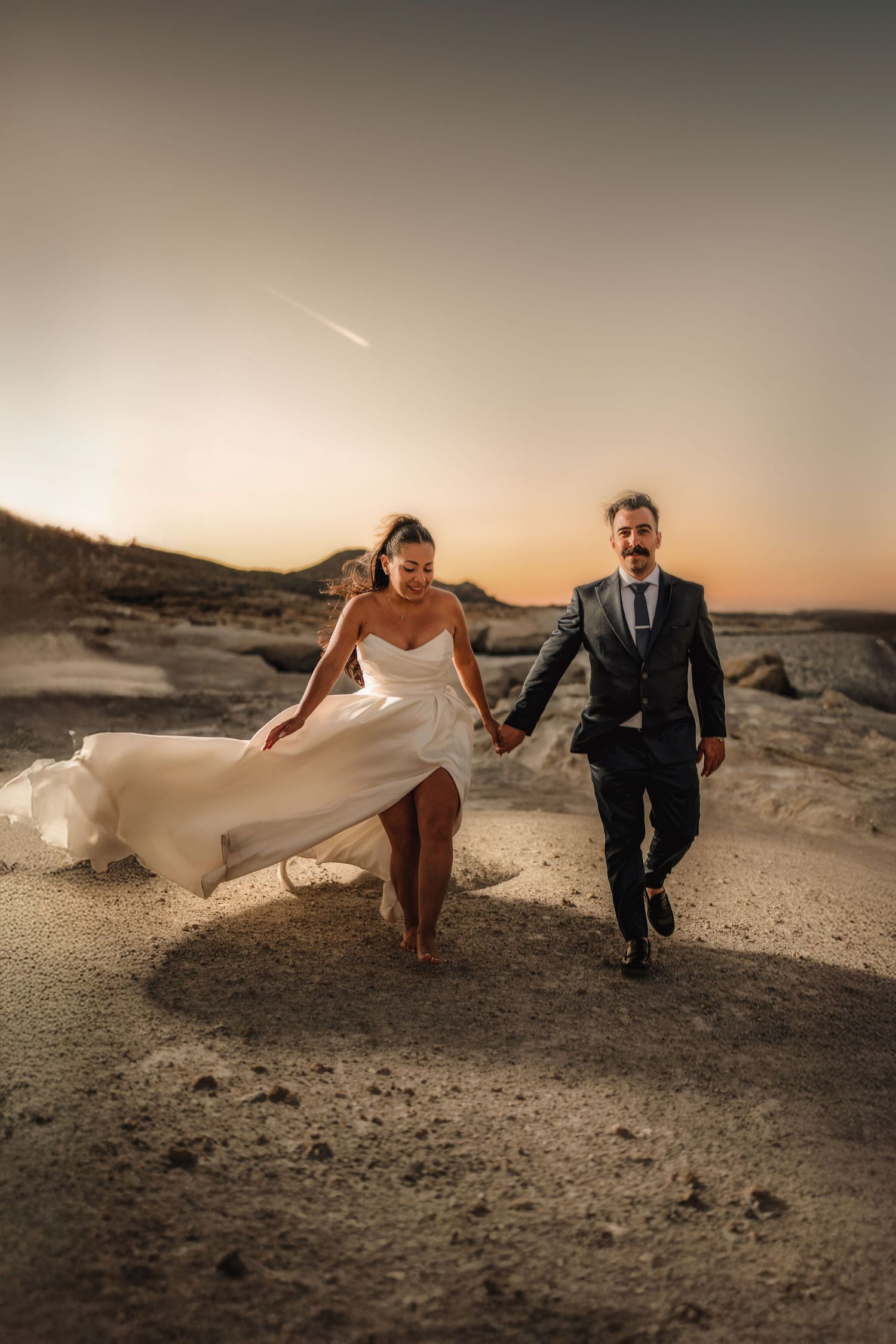 Couple in wedding attire running playfully across a desert landscape at sunset.