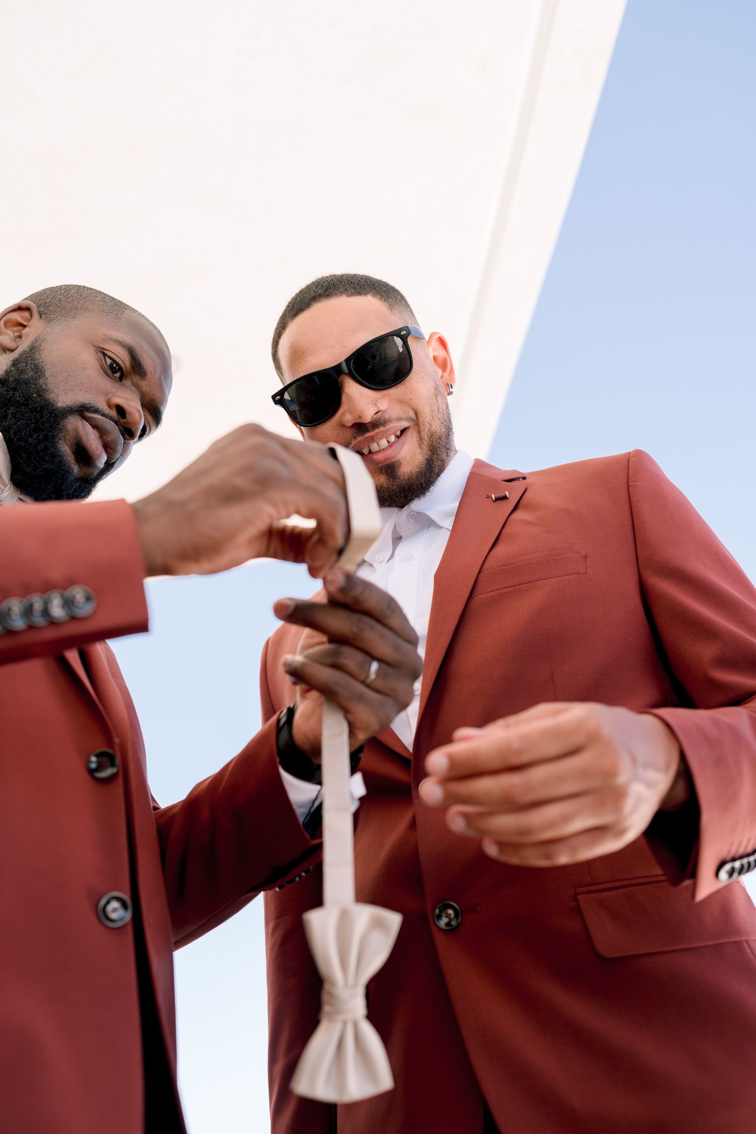 Two people in burgundy suits adjust bow ties while getting ready for a formal event.