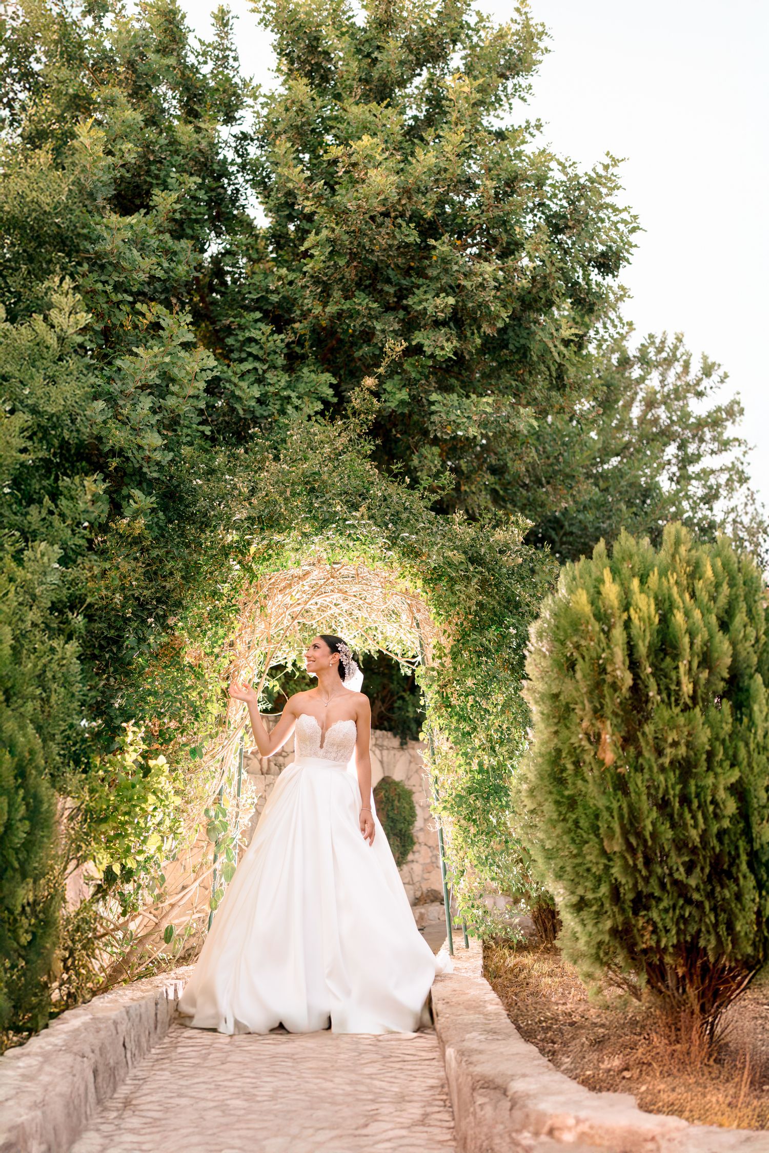 Bride in white gown walks through a floral-covered garden archway.