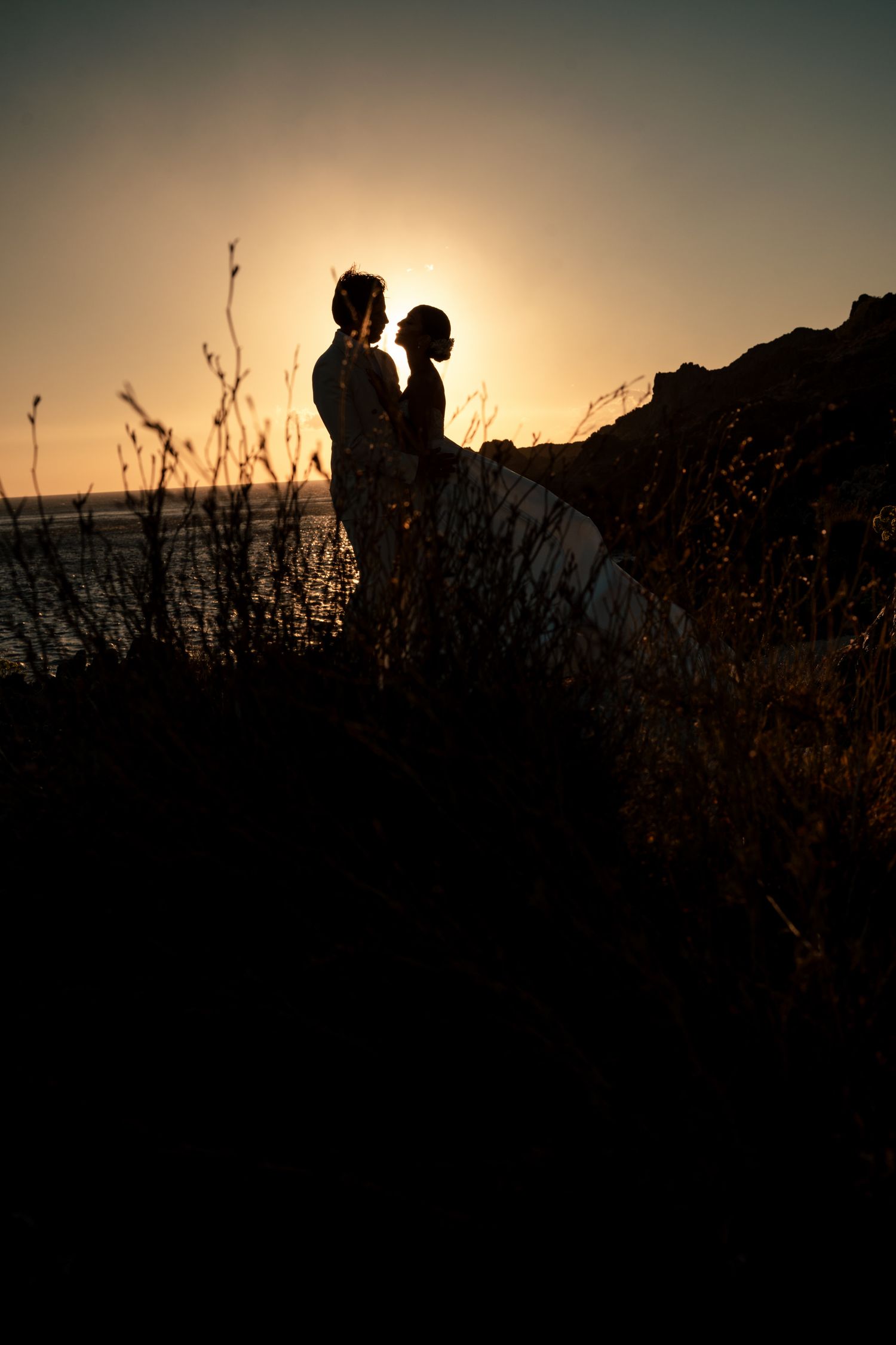 Silhouettes of a couple against a dramatic sunset sky with coastal vegetation.