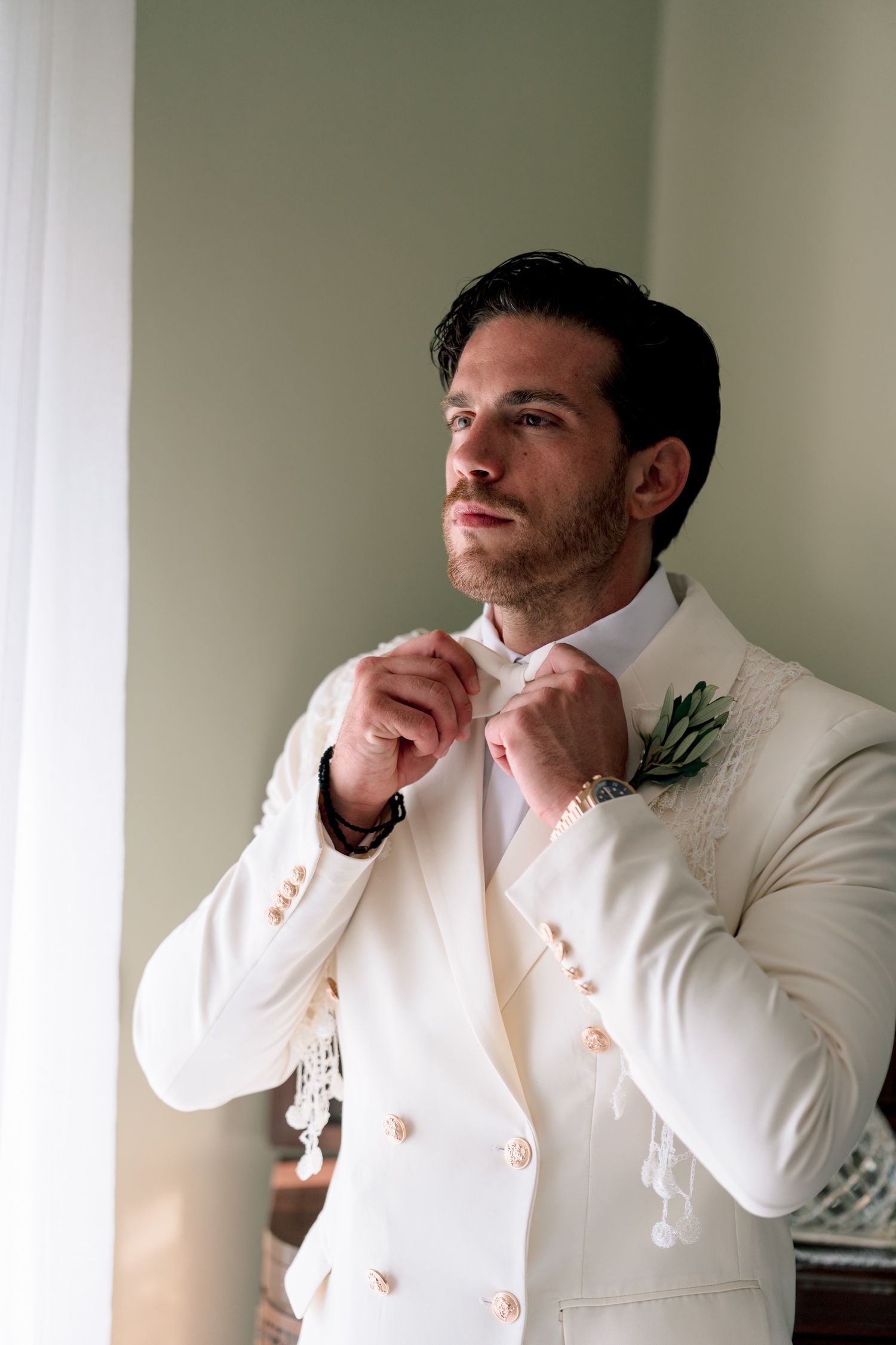 A groom in a white tuxedo jacket adjusts his collar while getting ready.
