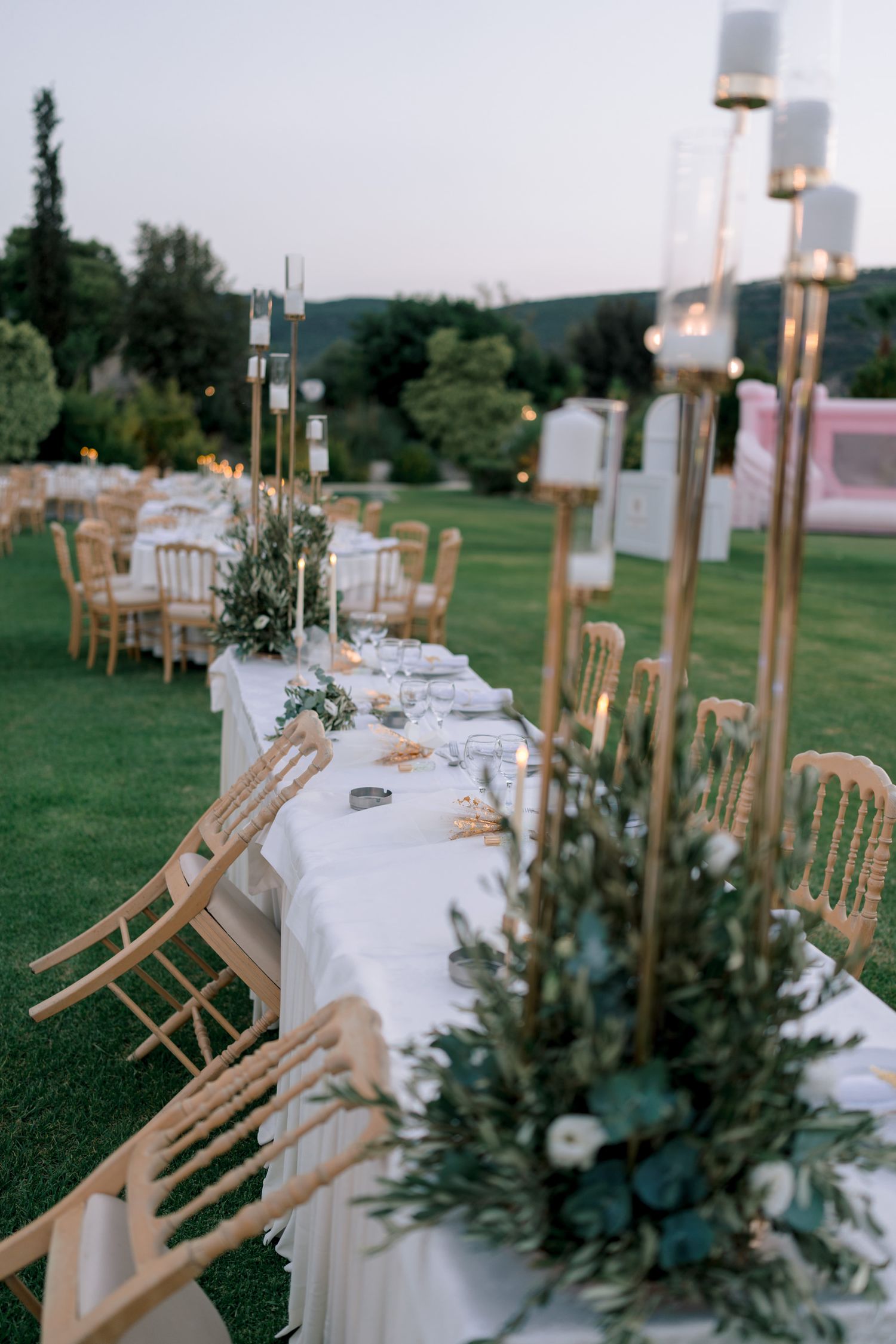Wedding reception tables decorated with tall candle holders and greenery centerpieces on white tablecloths.