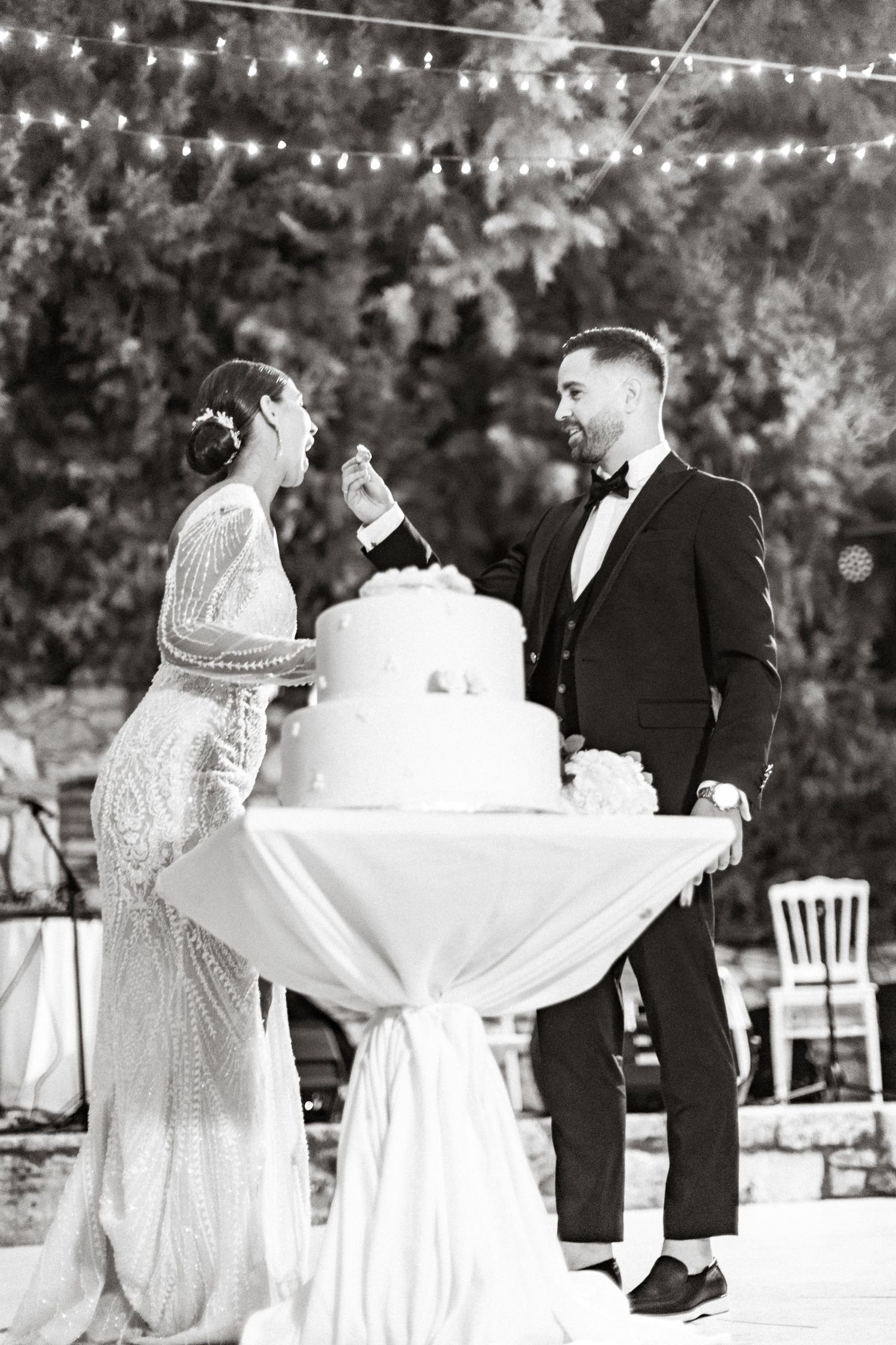 Wedding cake and couple toasting during an outdoor evening reception with string lights.