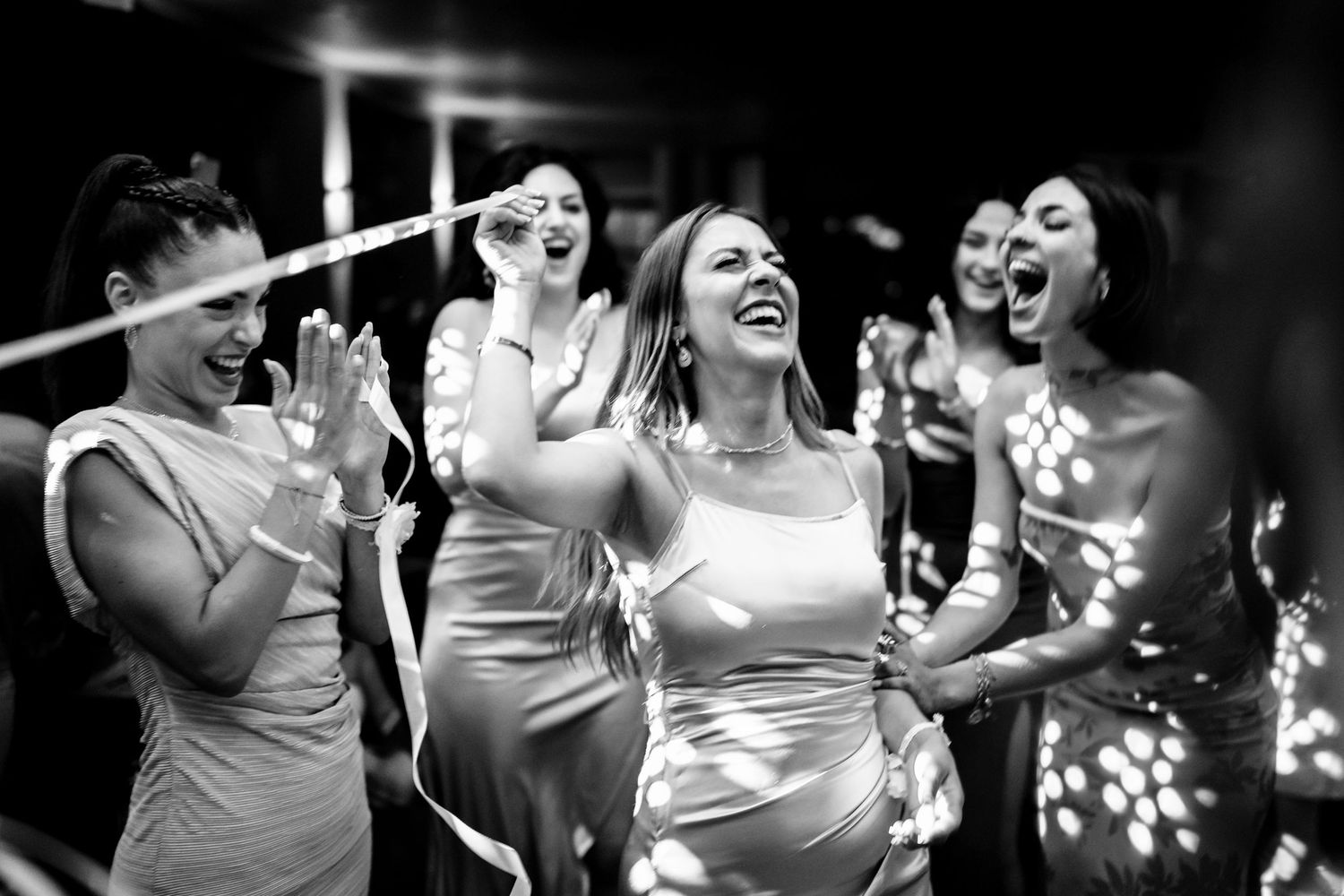 Wedding party members dance energetically in black and white photo with dramatic lighting.
