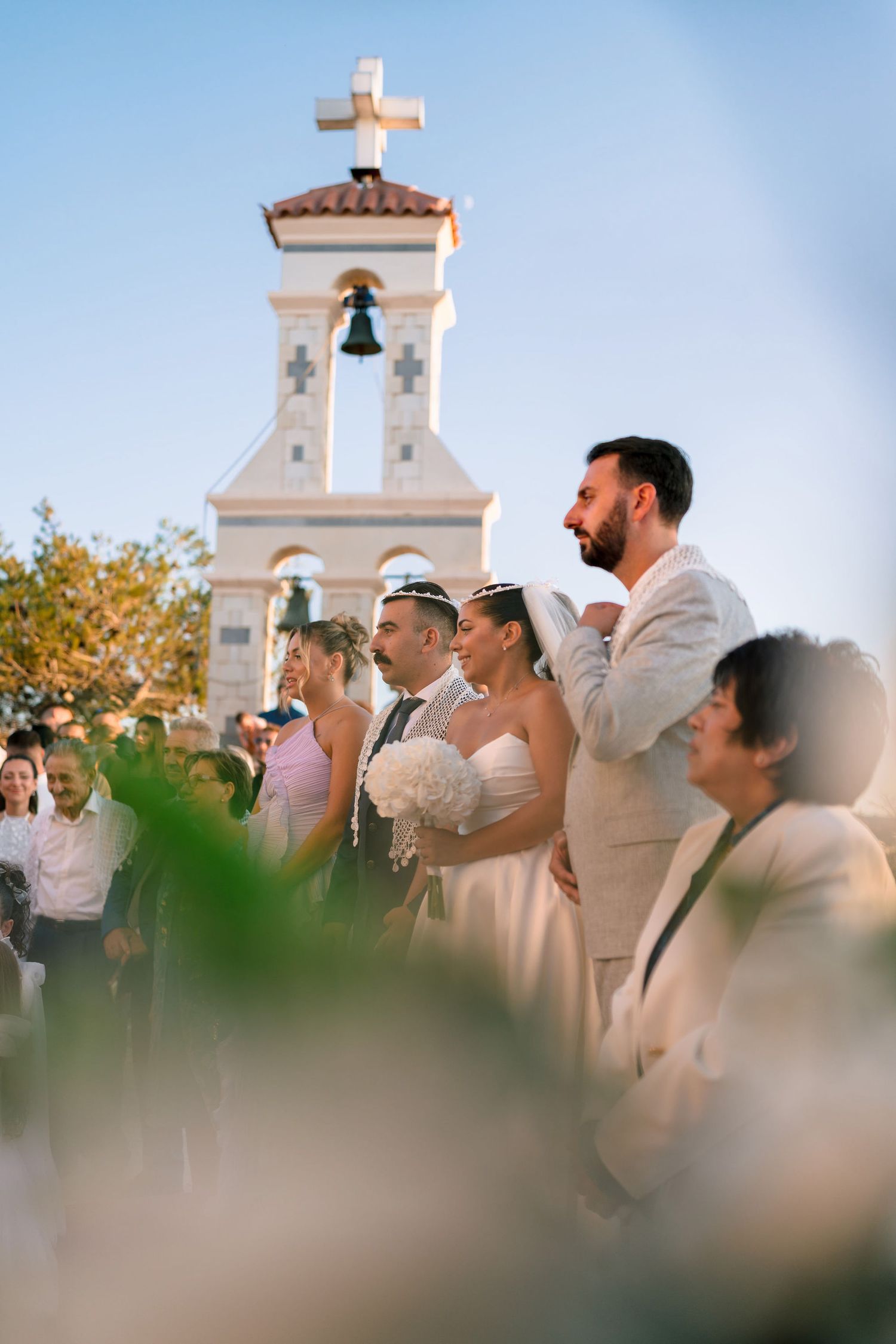 Wedding party standing in front of a white church bell tower during an outdoor ceremony.