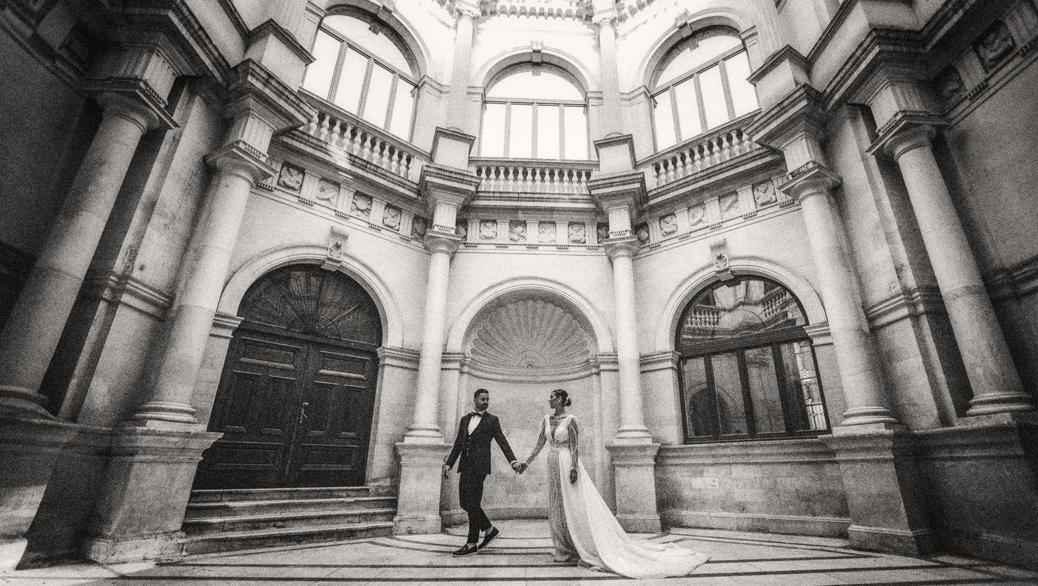 Wedding couple silhouetted in grand architectural rotunda with ornate columns and arched doorways.