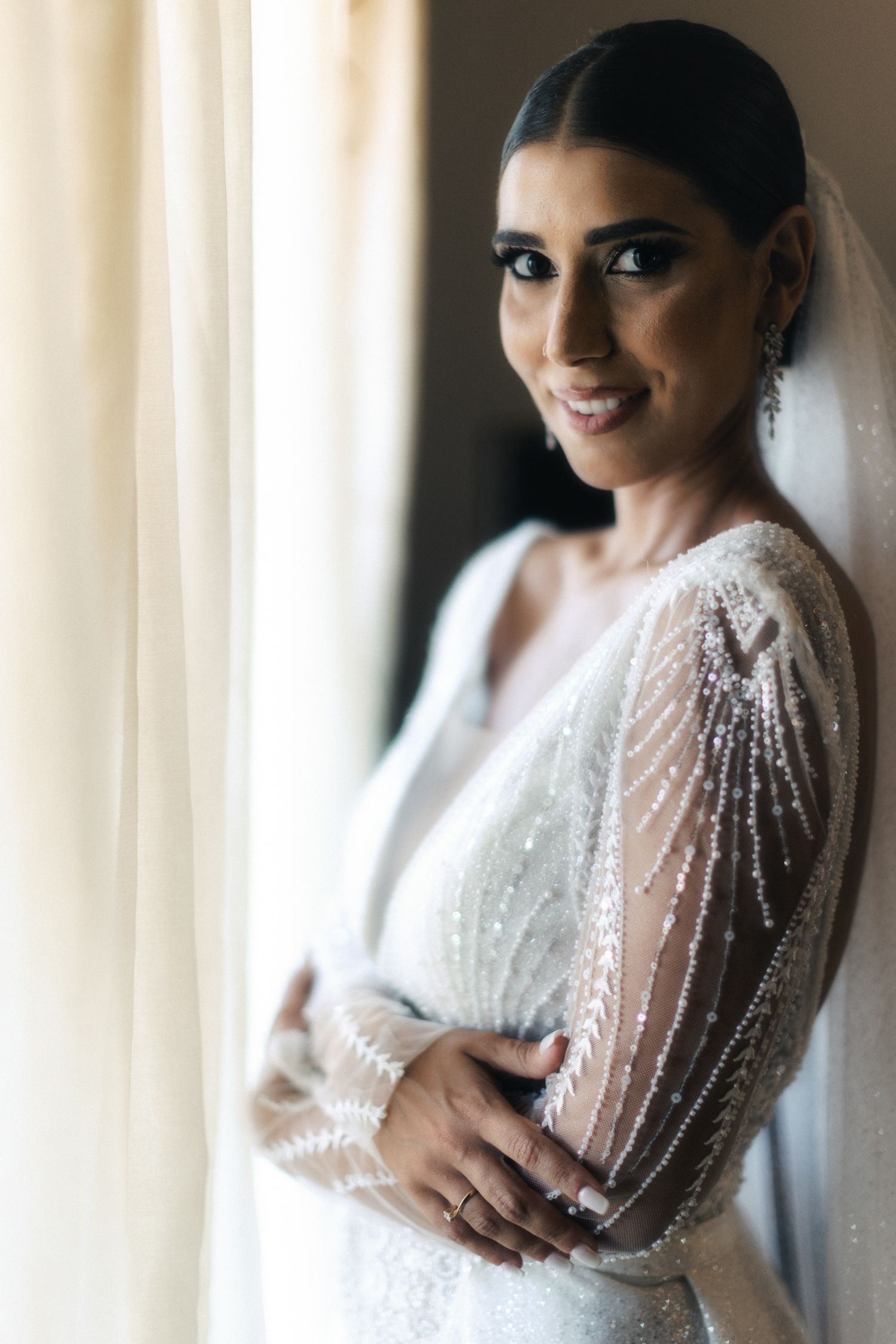 Elegant bride in a sparkly long-sleeve wedding gown poses by window light.