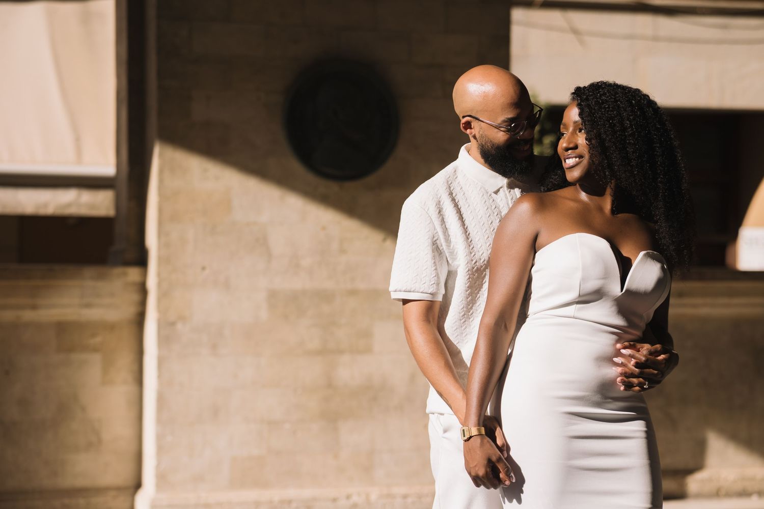 Couple embracing in white outfits against a textured wall with dramatic palm tree shadows.