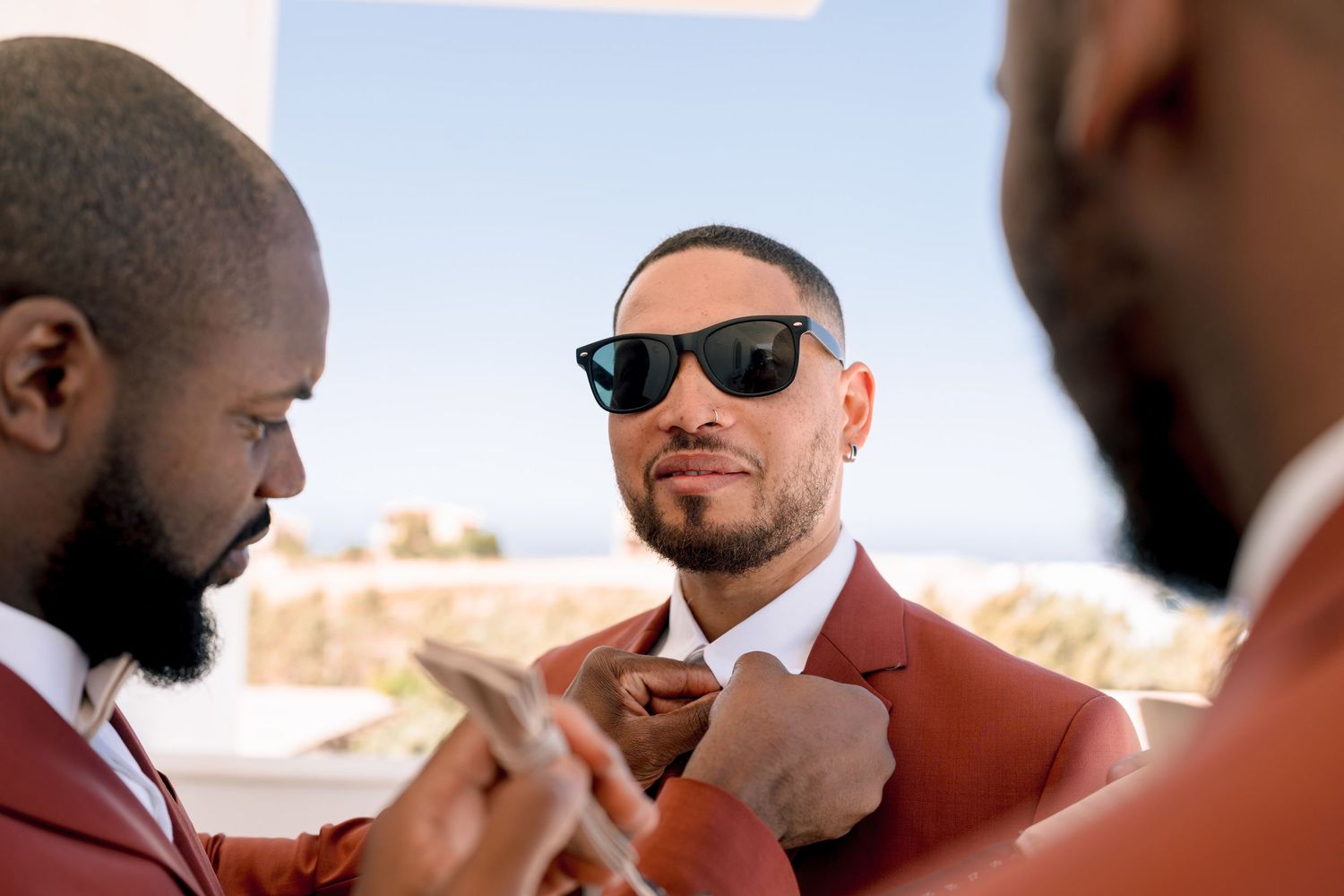 Wedding party members in burgundy suits and sunglasses help with formal attire preparations.