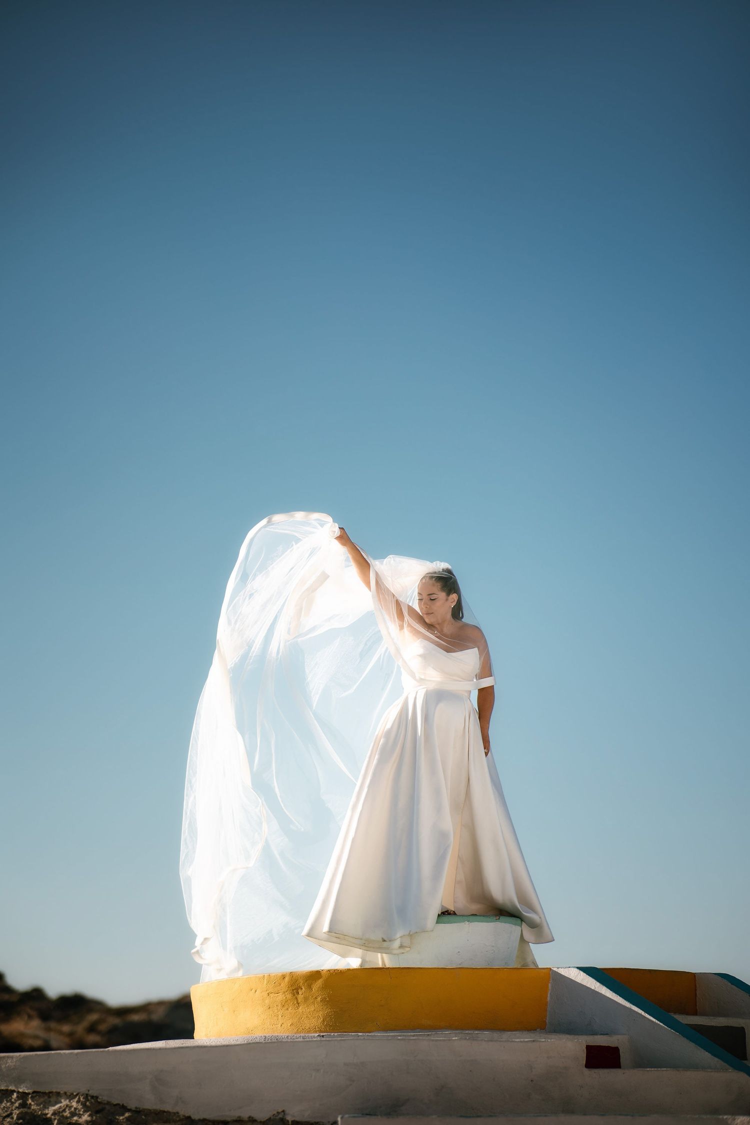 Artistic wedding photography with flowing white dress against bright blue sky.