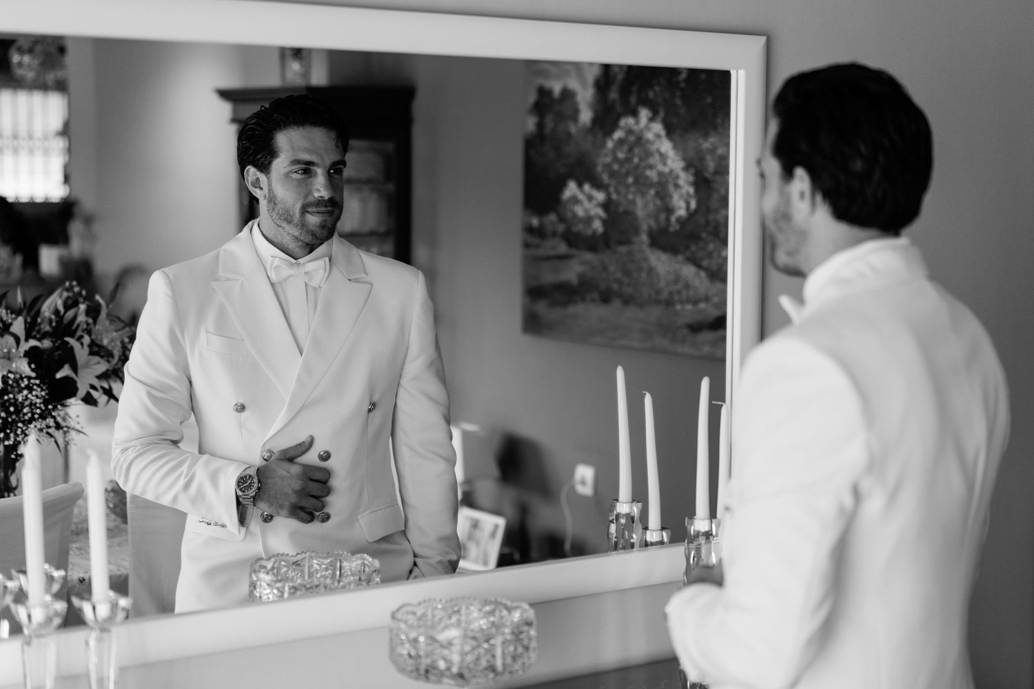 Reflection of a groom in formal white attire looking in a mirror with candles on the dresser.