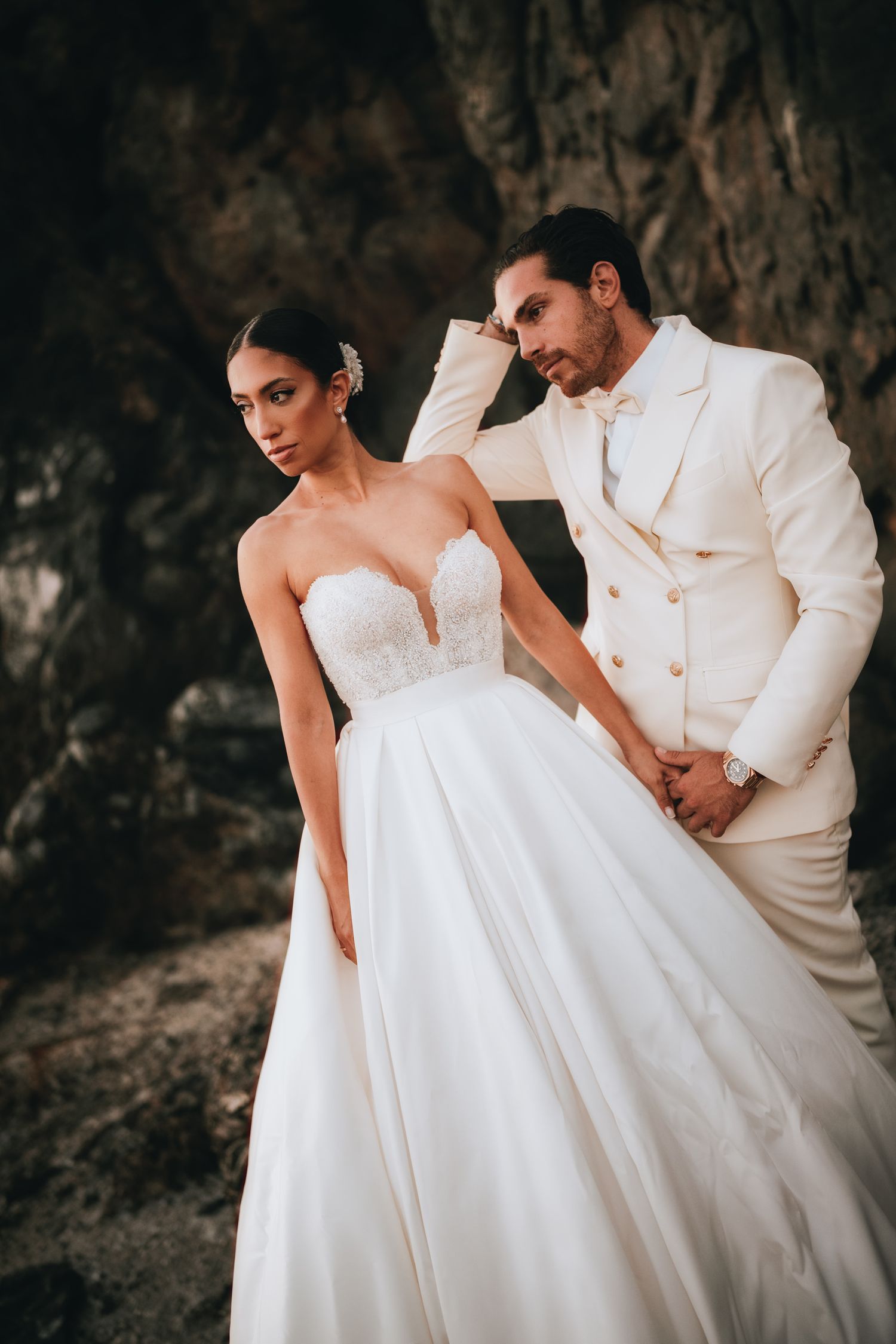 Romantic wedding photos featuring couple in white formal wear posing against textured stone wall.