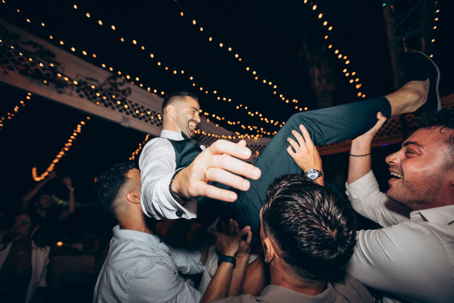 Wedding guests dancing and celebrating under warm string lights at an evening reception.