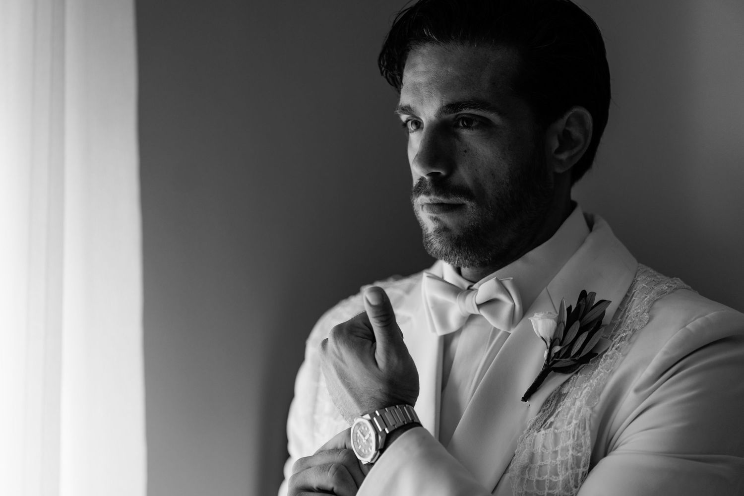 Black and white photo of groom in white tuxedo making final preparations.