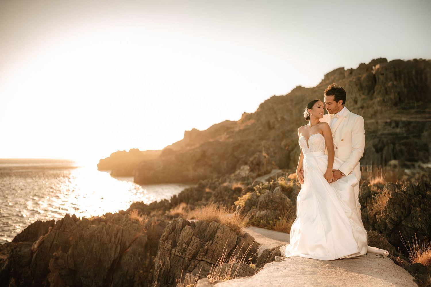 Couple in wedding attire embracing on coastal cliff at sunset.