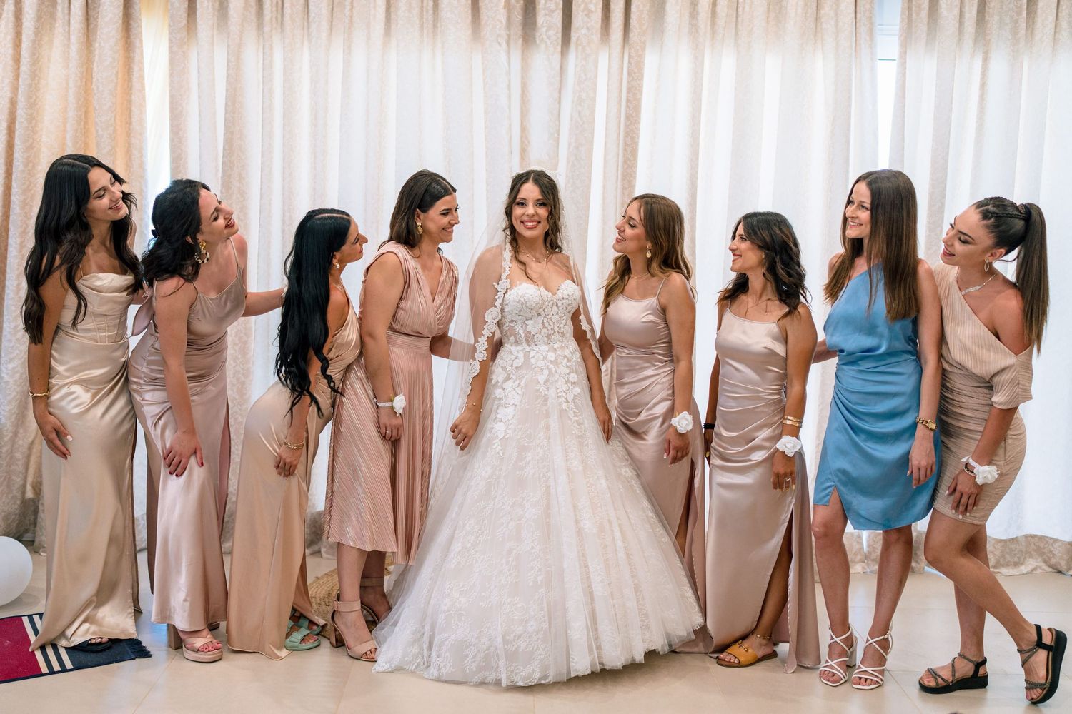 Bridesmaids in neutral-toned dresses gather around bride wearing white ballgown in elegant indoor setting.