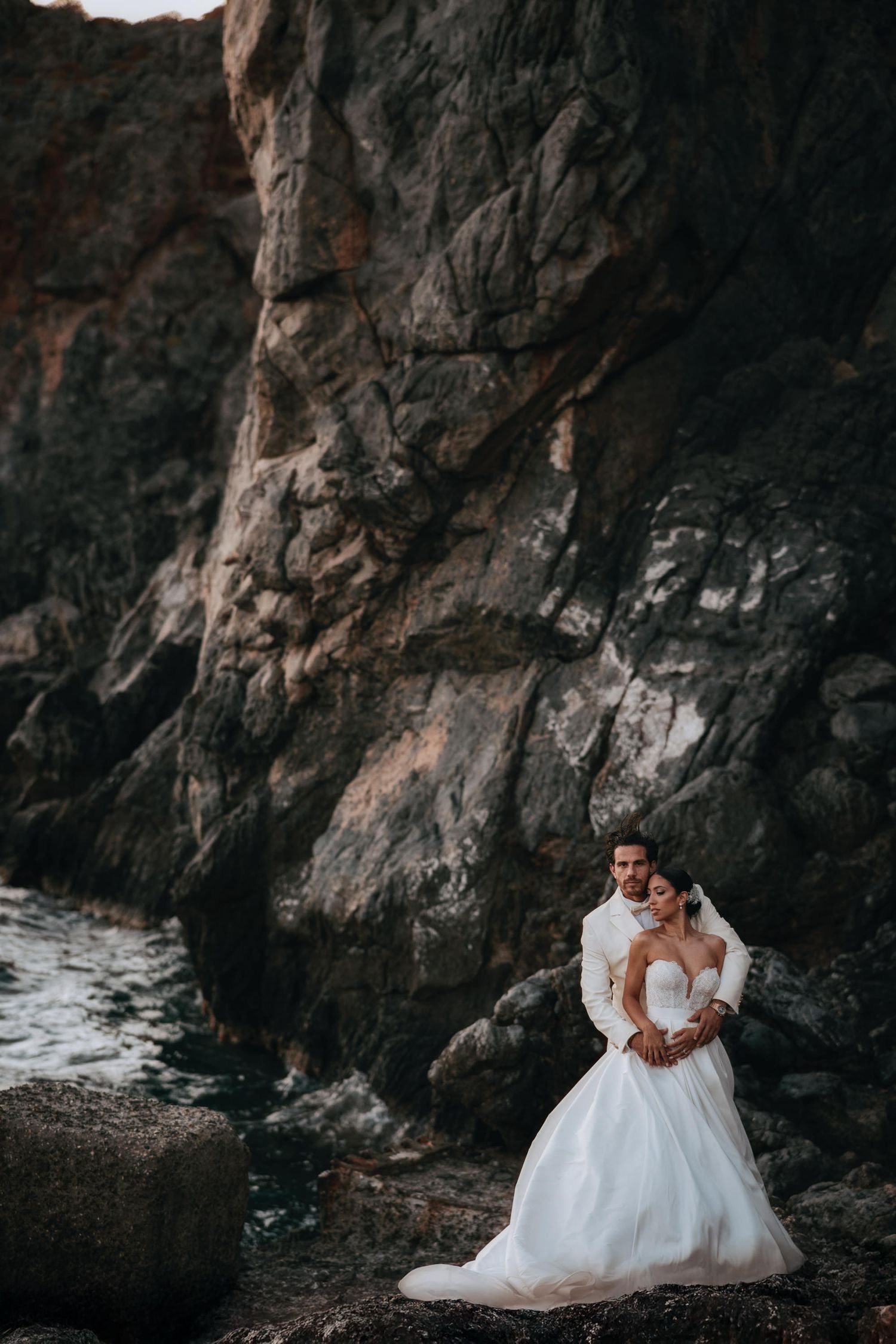 A wedding couple embraces near dramatic coastal rock formations.