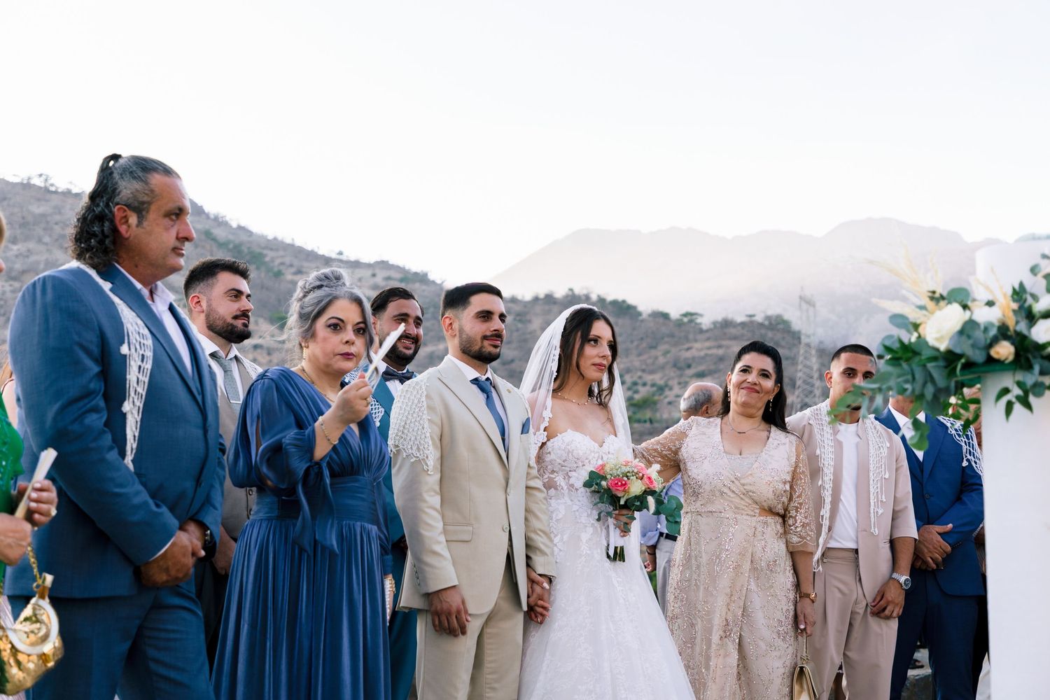 Wedding party in coordinated blue and beige attire poses against mountain backdrop at outdoor celebration.