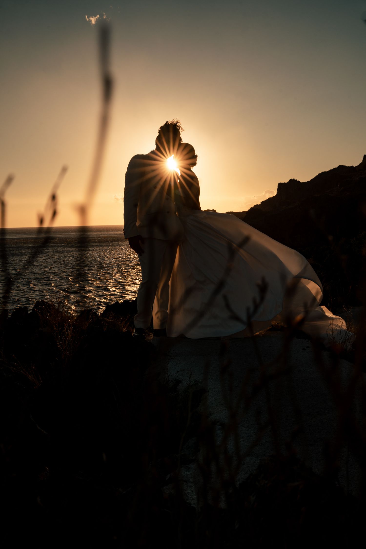 Silhouetted wedding couple against a setting sun over ocean waters.