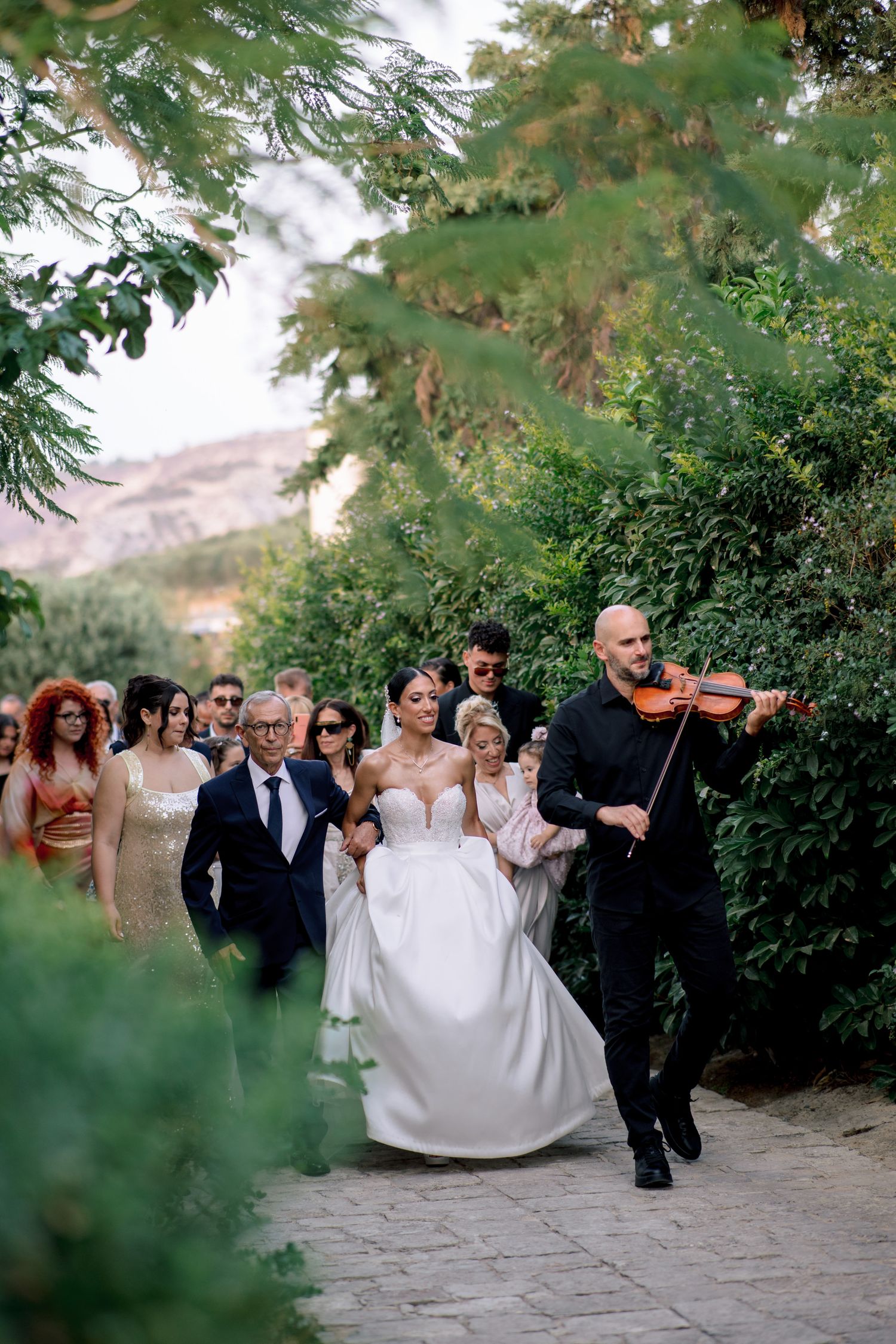 Wedding party walking along a garden path with a violinist playing music.