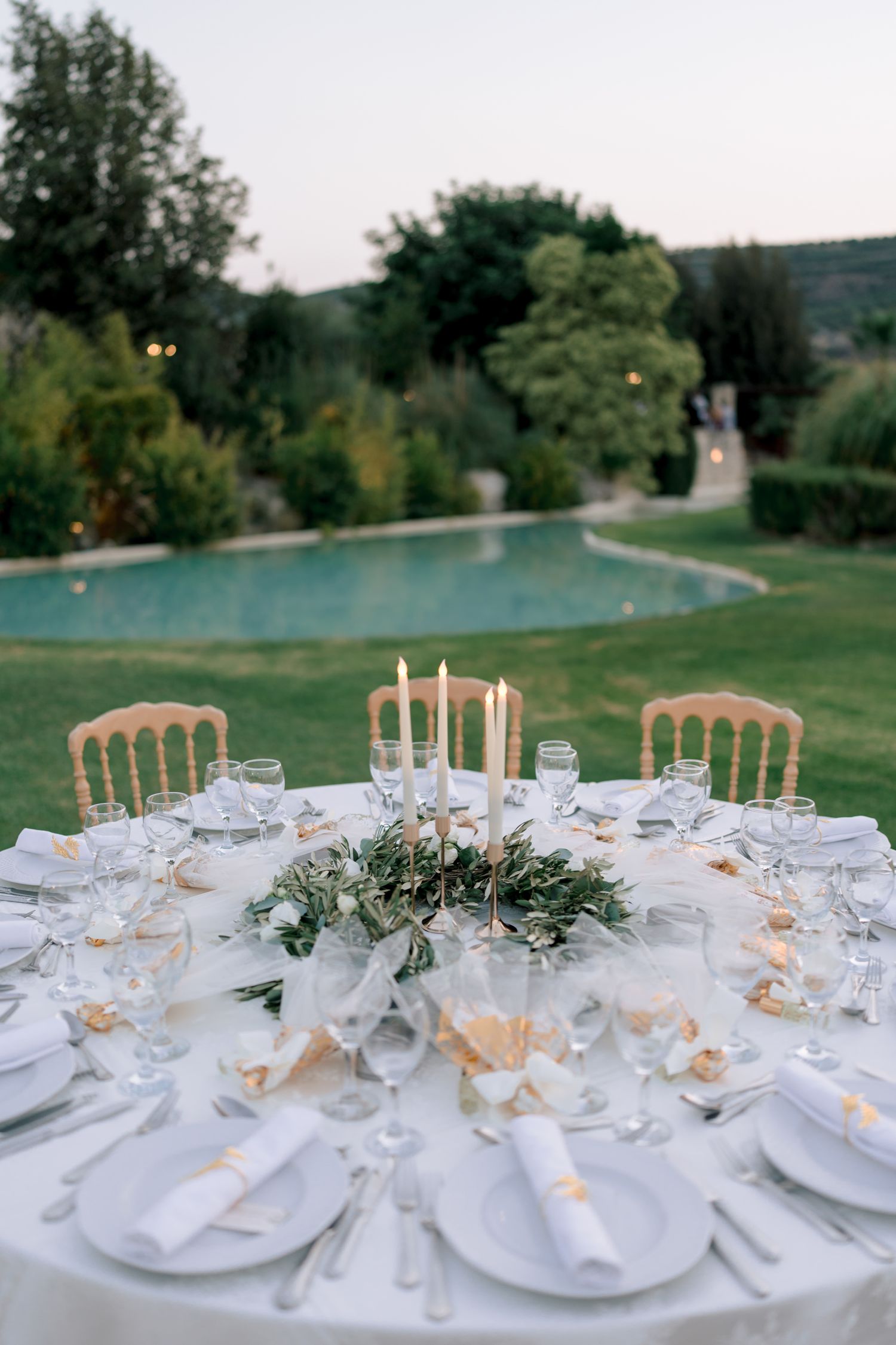 Elegant wedding reception tables set with white linens, glassware and golden chairs in an outdoor garden setting.
