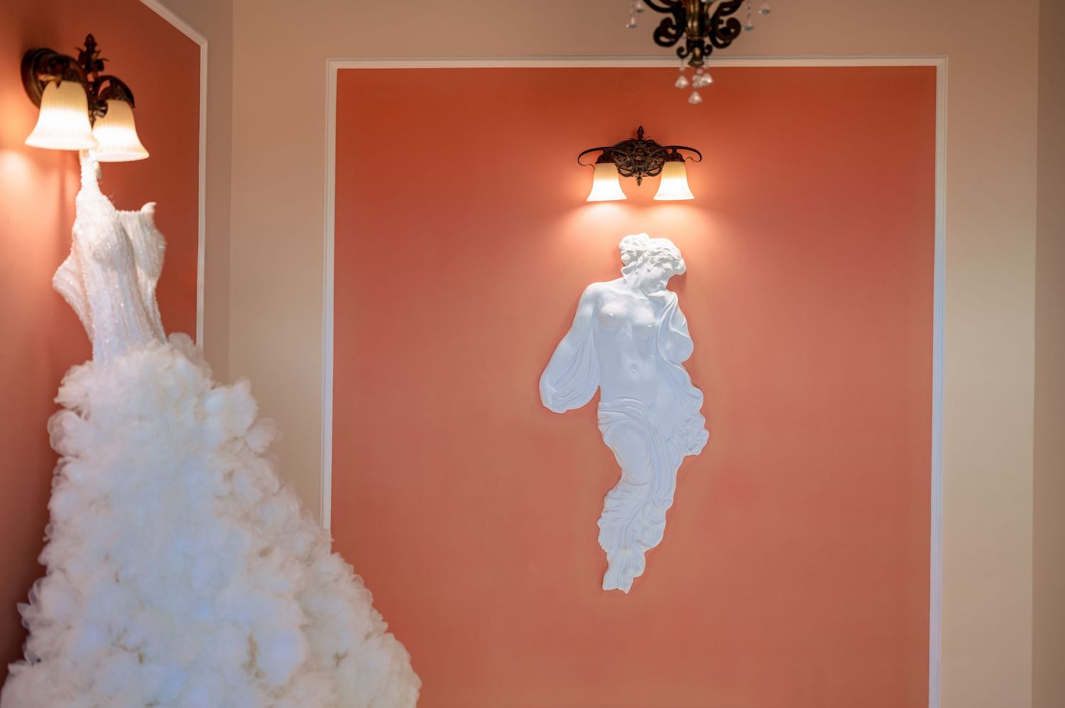 Wedding dress with dramatic tulle skirt displayed against coral-colored walls with lighting fixtures.