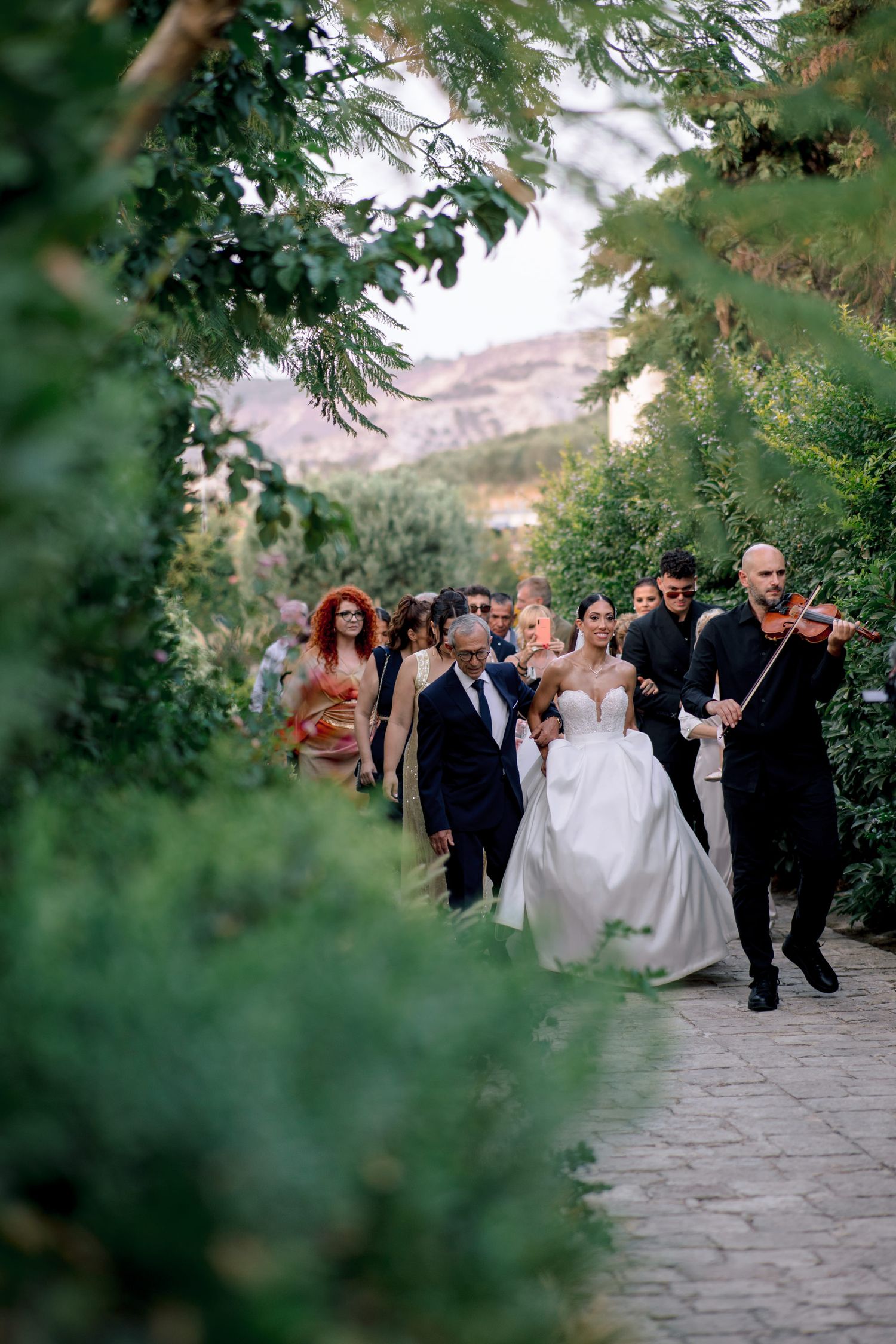 Wedding processional walking through lush garden path with violinist playing.