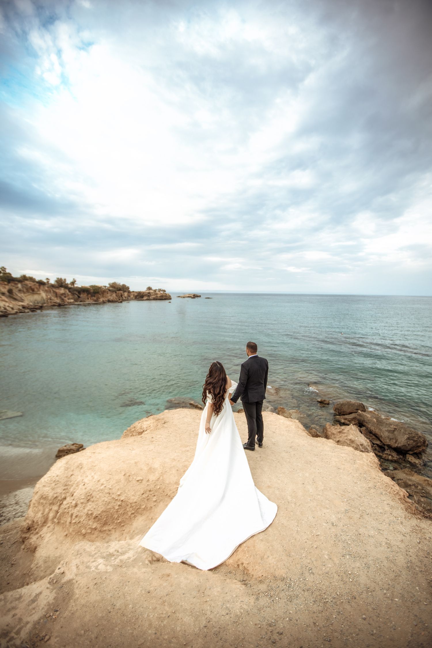 Newlyweds stand on coastal cliff overlooking turquoise waters under dramatic sky.