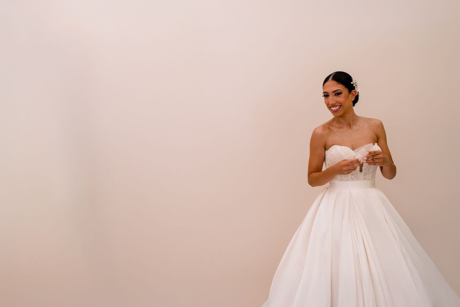 A bride in a white strapless wedding gown adjusts her dress in front of a neutral background.