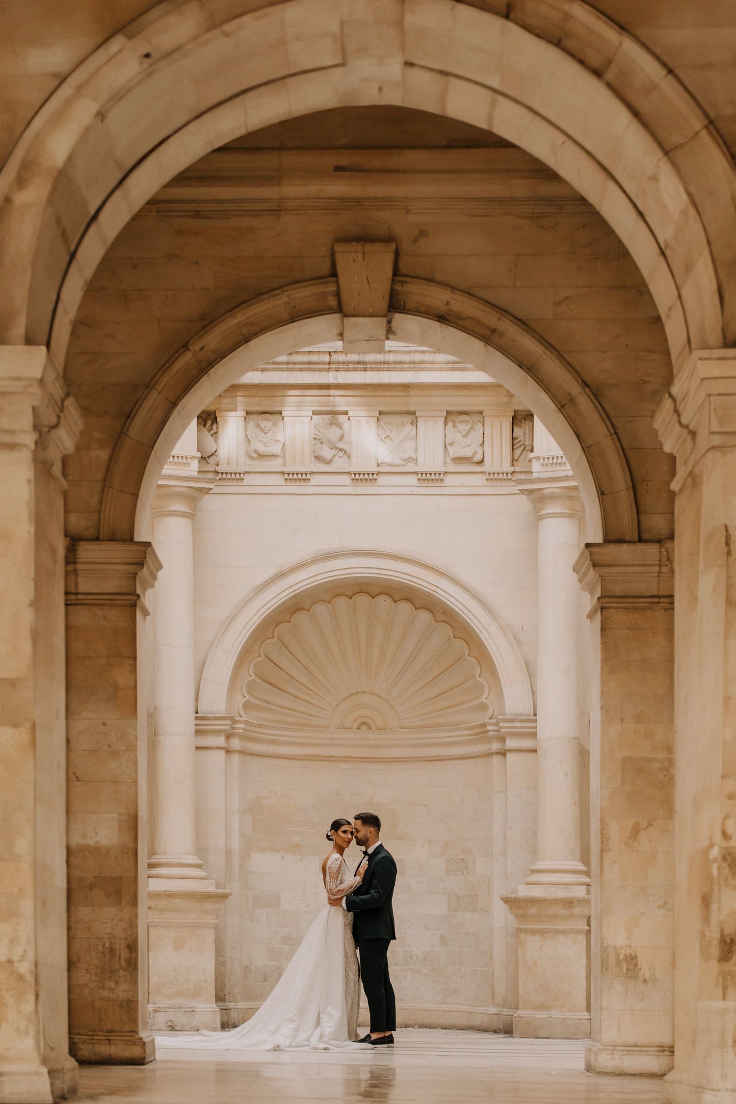 Wedding couple embracing under ornate stone archway with classical architecture.