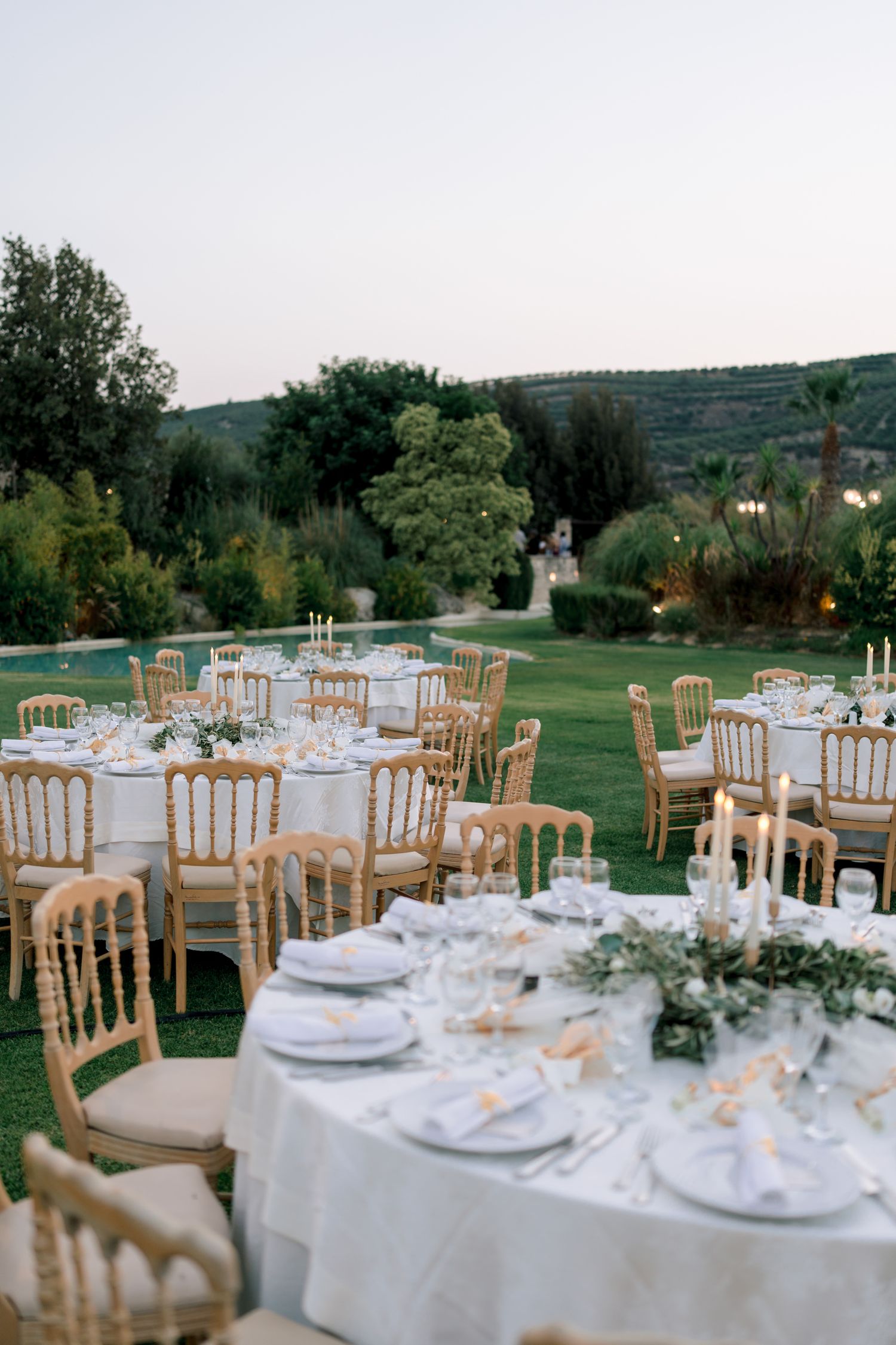 Elegant outdoor wedding reception setup with white tablecloths and gold chiavari chairs on a lush green lawn.