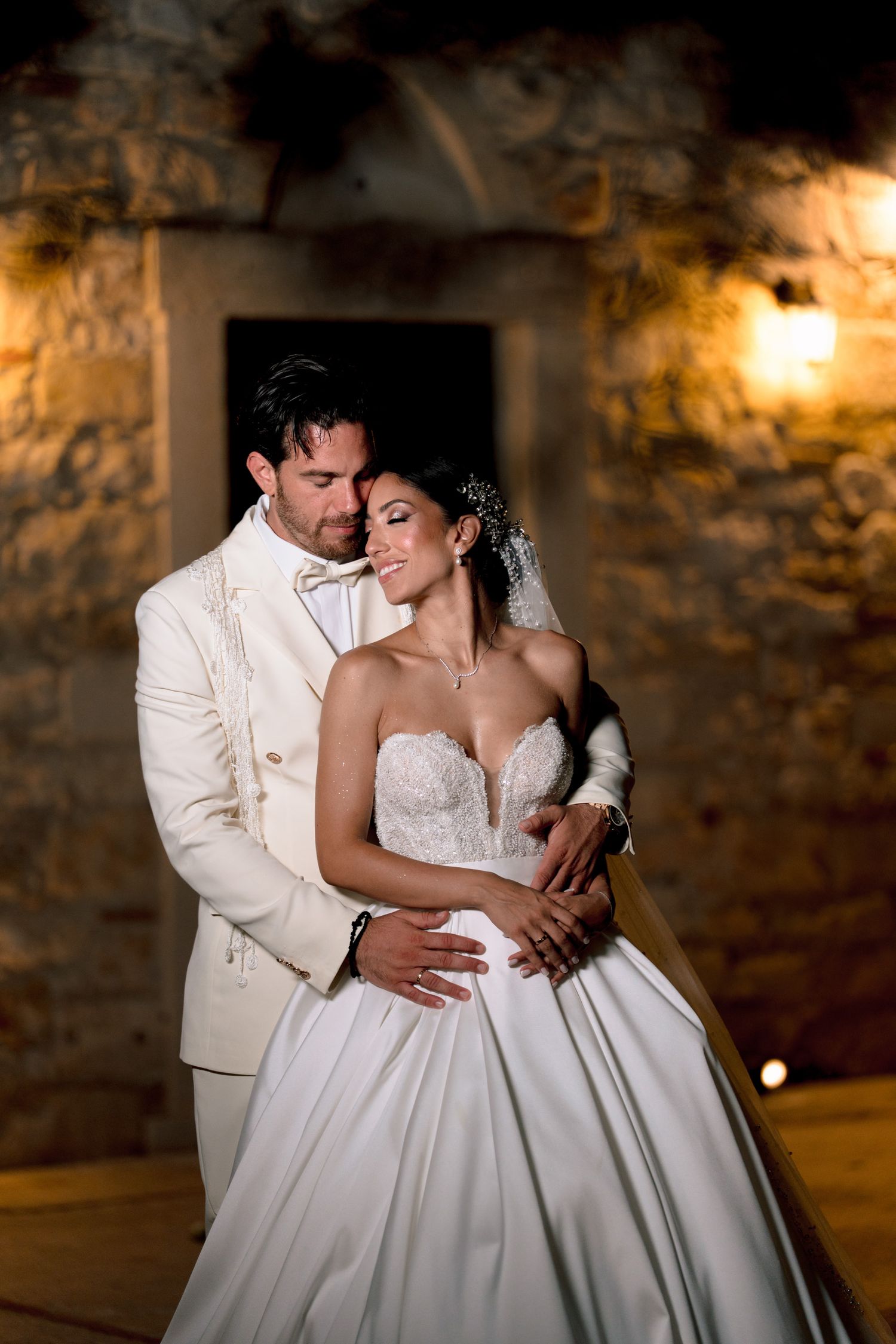 A tender moment between newlyweds in white attire against a rustic stone wall.