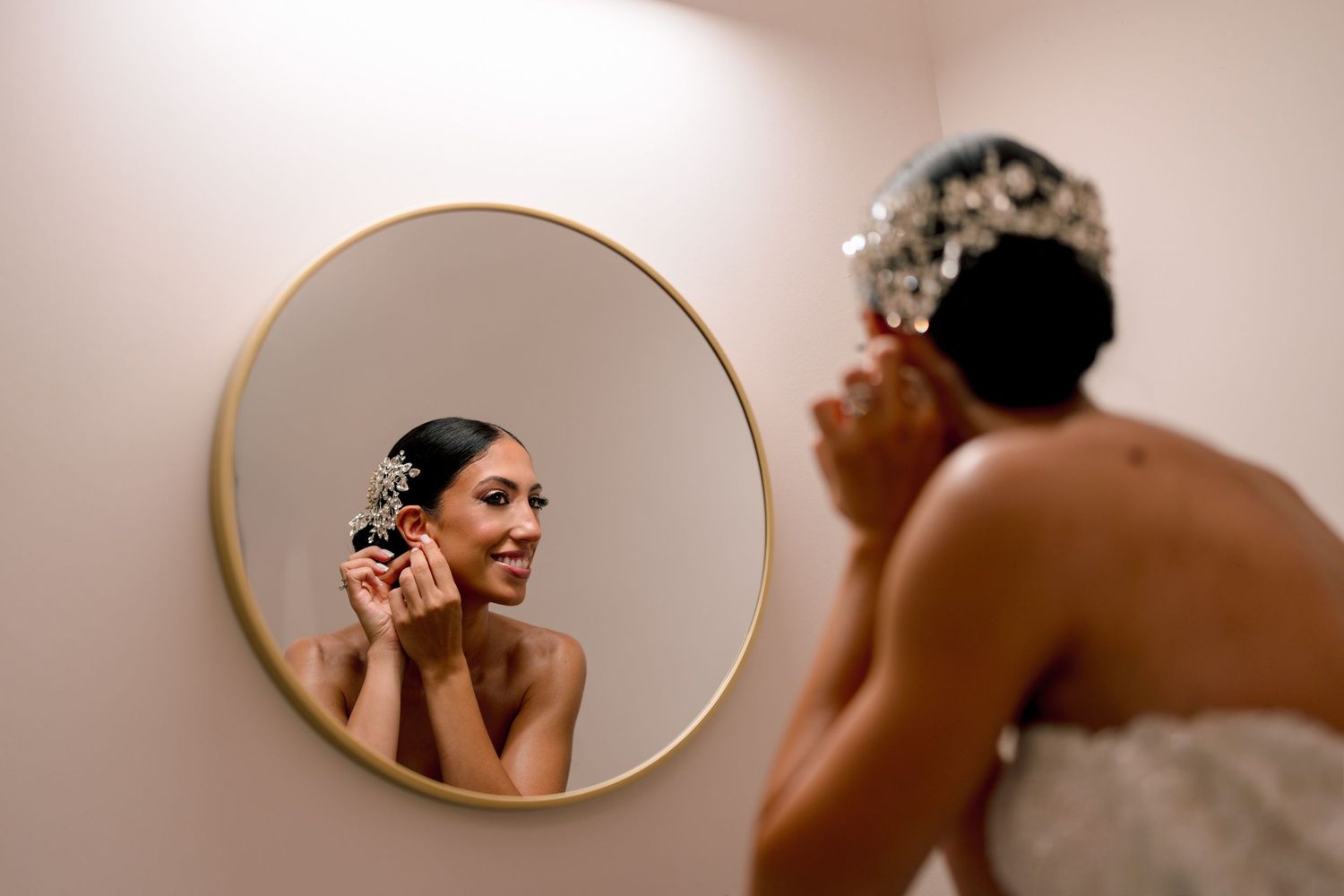 Bride getting ready in front of oval mirror with warm lighting and elegant decor.