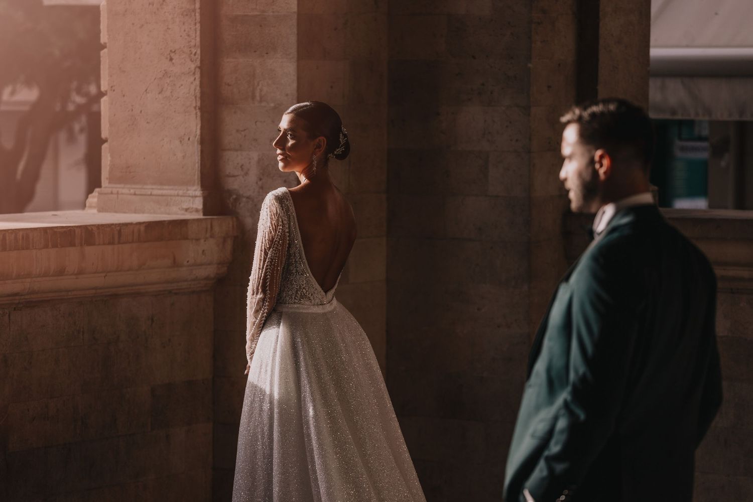 A bride in a lace dress stands in warm sunlight while her partner looks on.