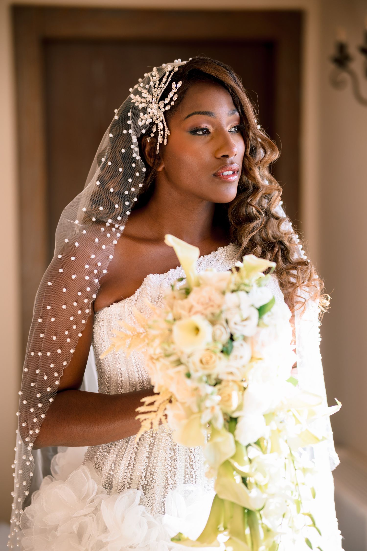 Bride wearing a pearl-dotted veil and holding a white and yellow bouquet in romantic lighting.