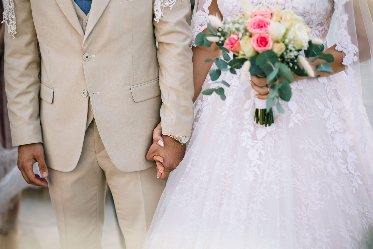 Wedding party in beige suits and white dress holding pink floral bouquets outdoors.
