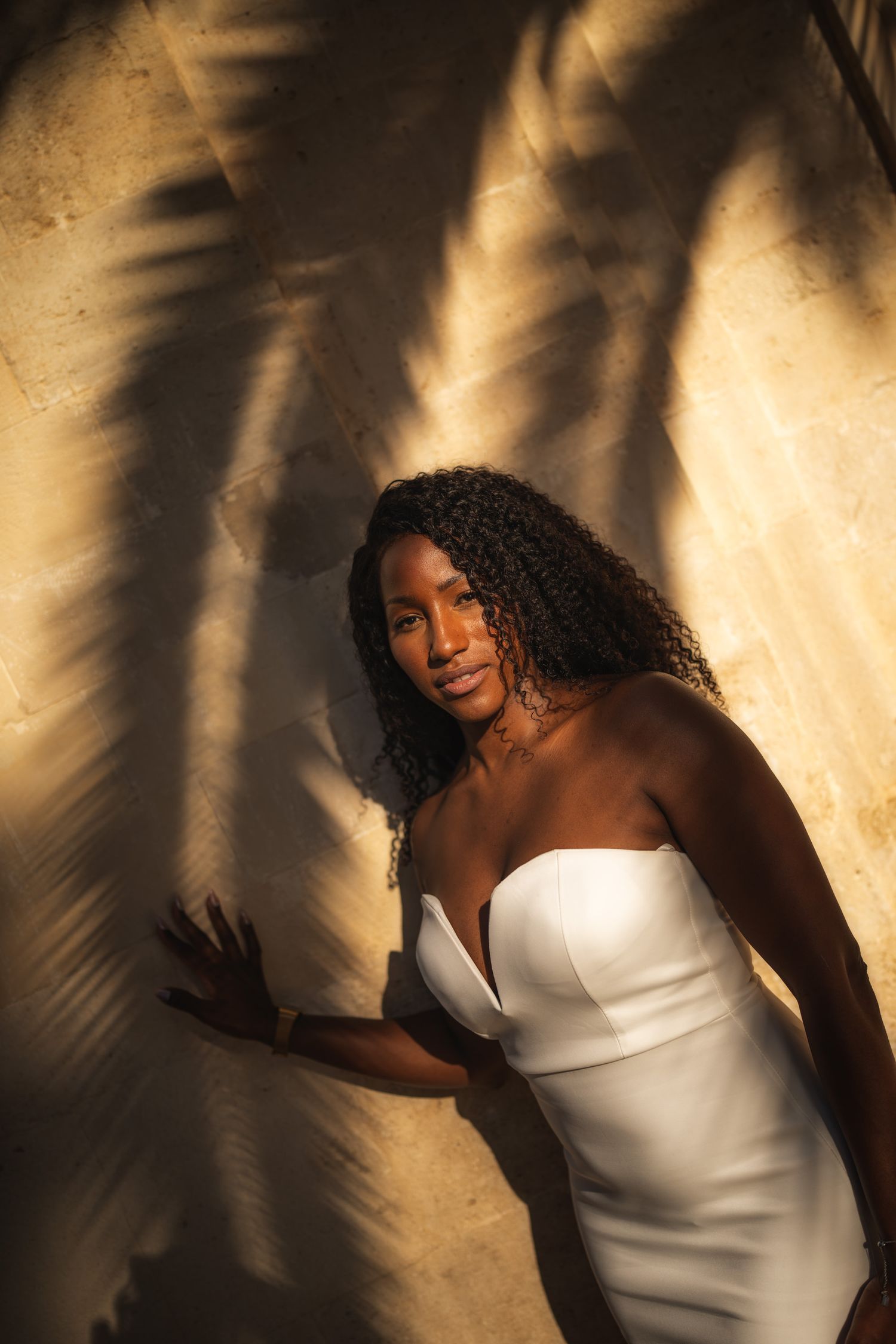 Woman in white dress poses elegantly with palm frond shadows creating dramatic lighting.