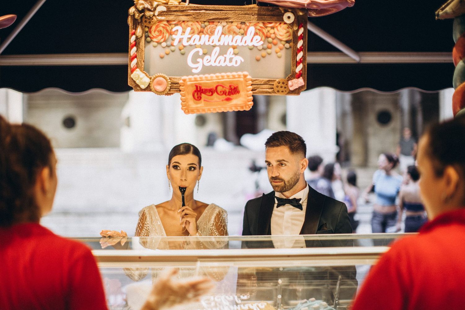 Couple in formal attire at gelato counter with servers in red uniforms.