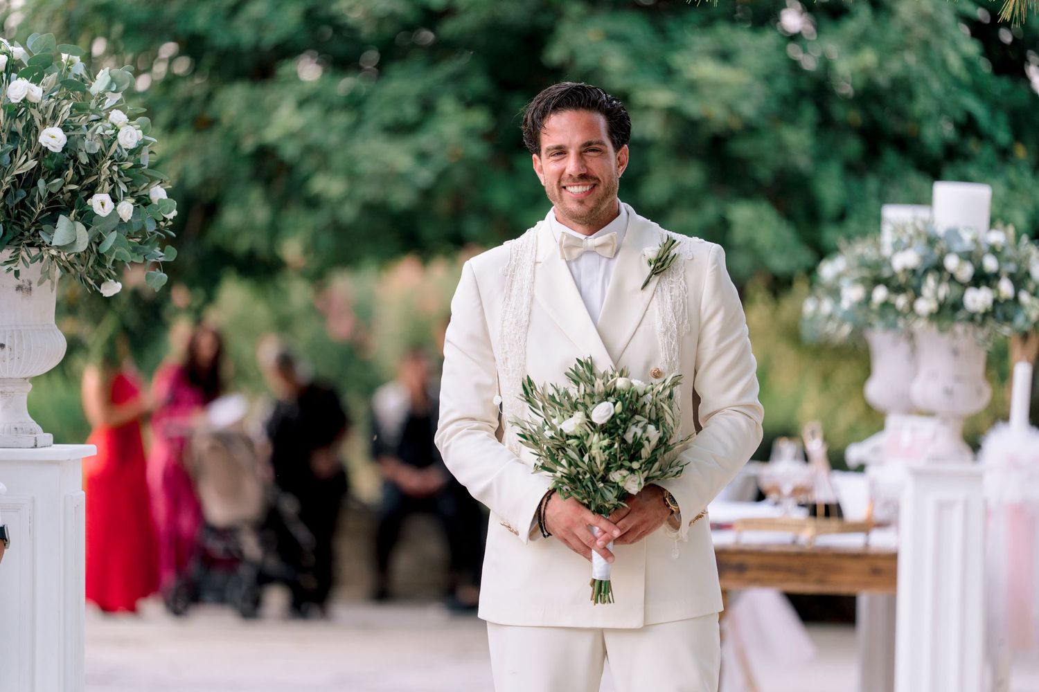 A groom in an all-white suit holding a bouquet of greenery.